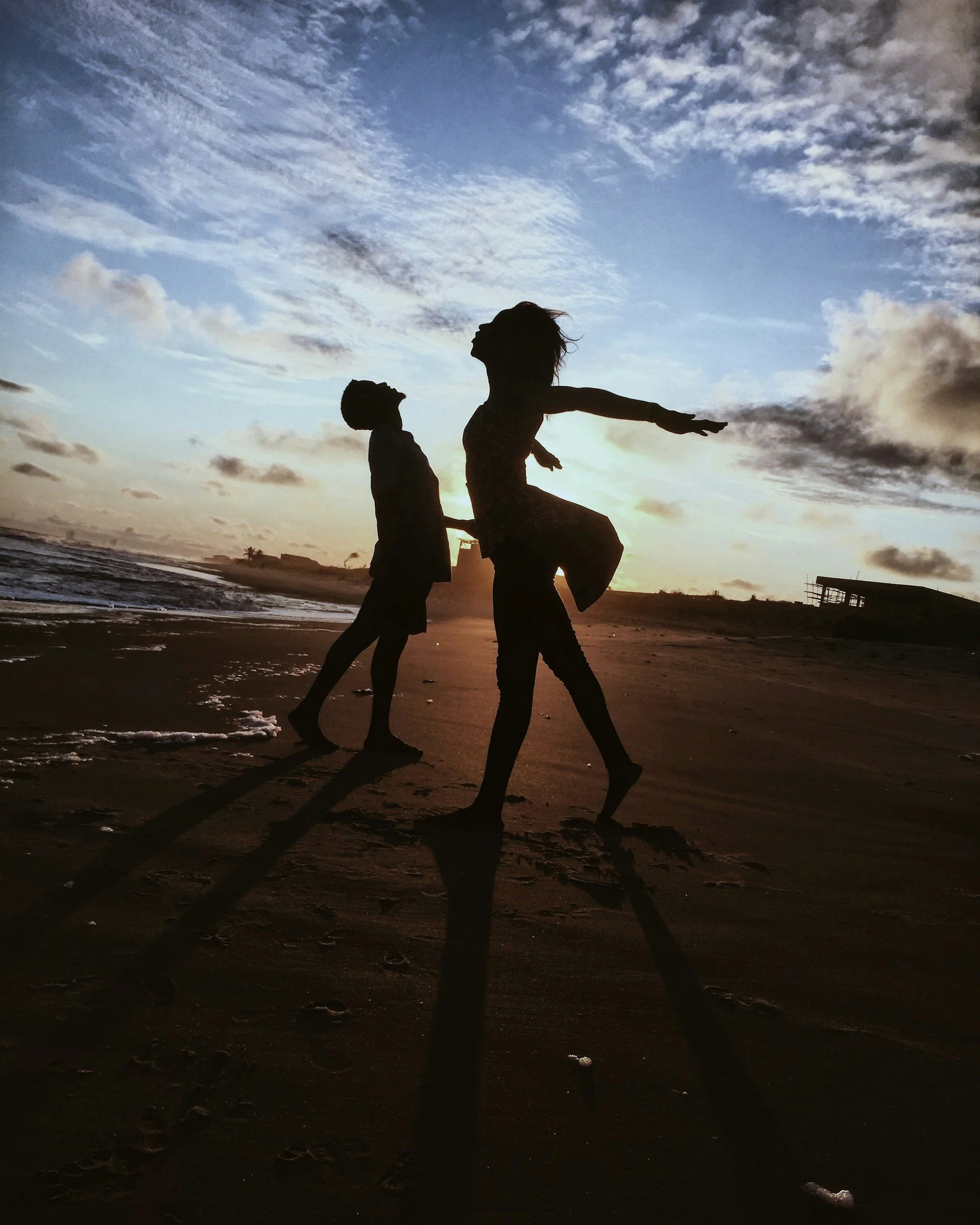 Silhouettes of three children playing on the beach during sunset, with the sky filled with clouds and reflections on the sand.