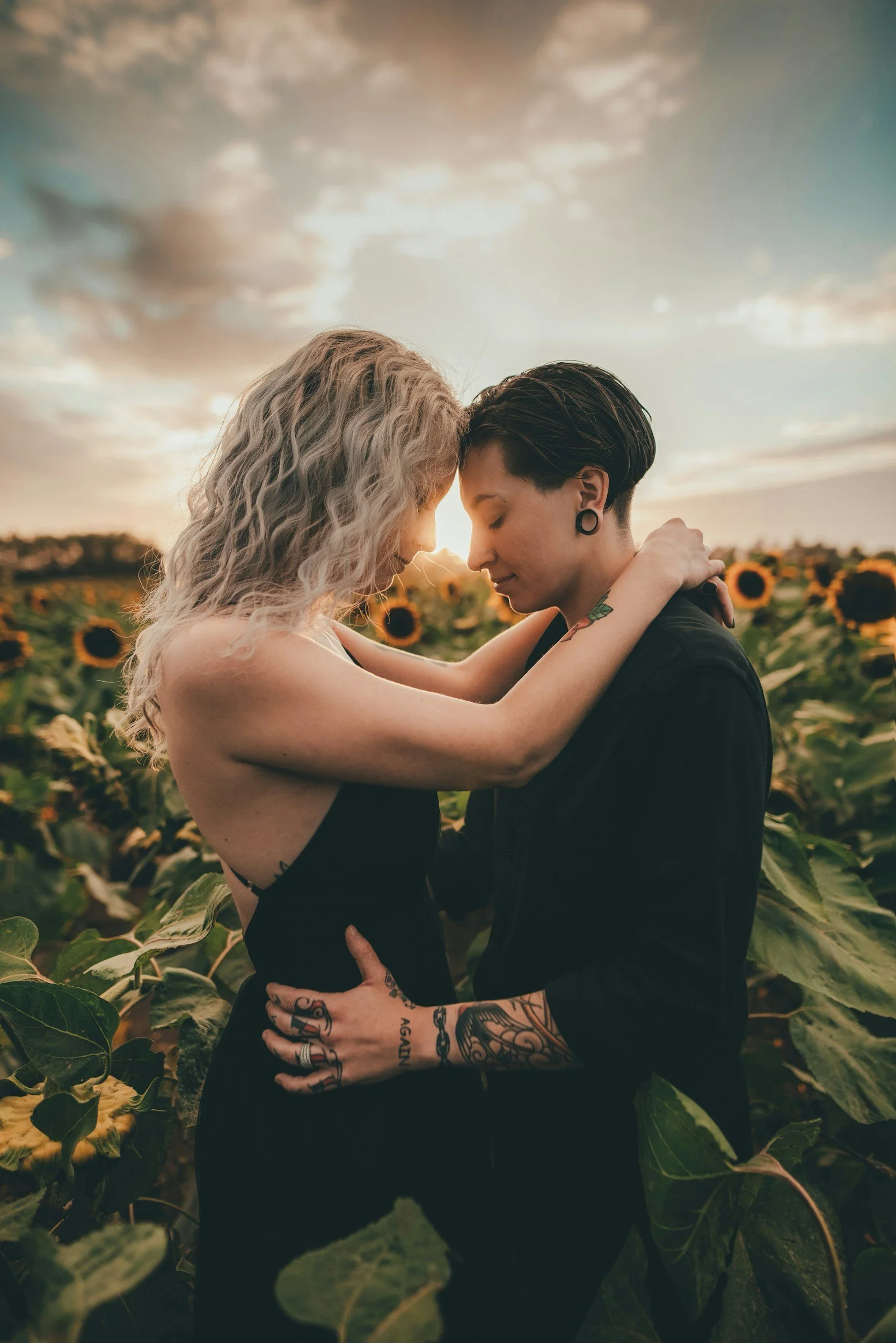 Two women embrace in a sunflower field at sunset, with their foreheads touching and their eyes closed.