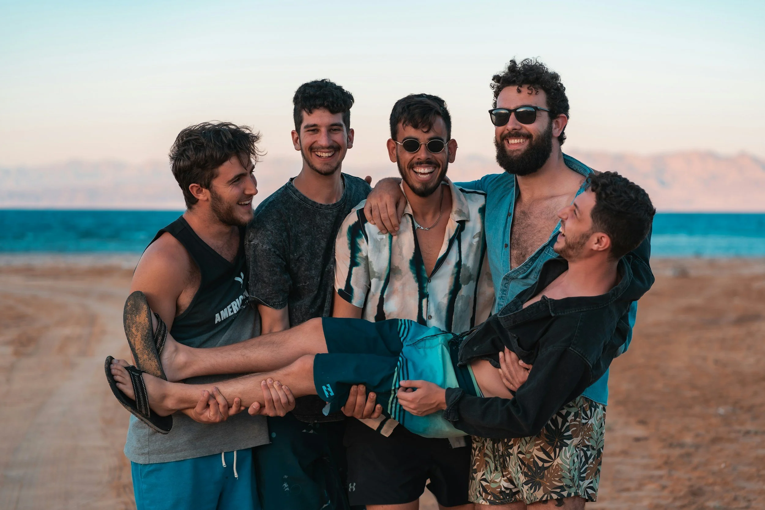 A group of six young men smiling and laughing together at the beach, with the ocean and a sunset in the background.