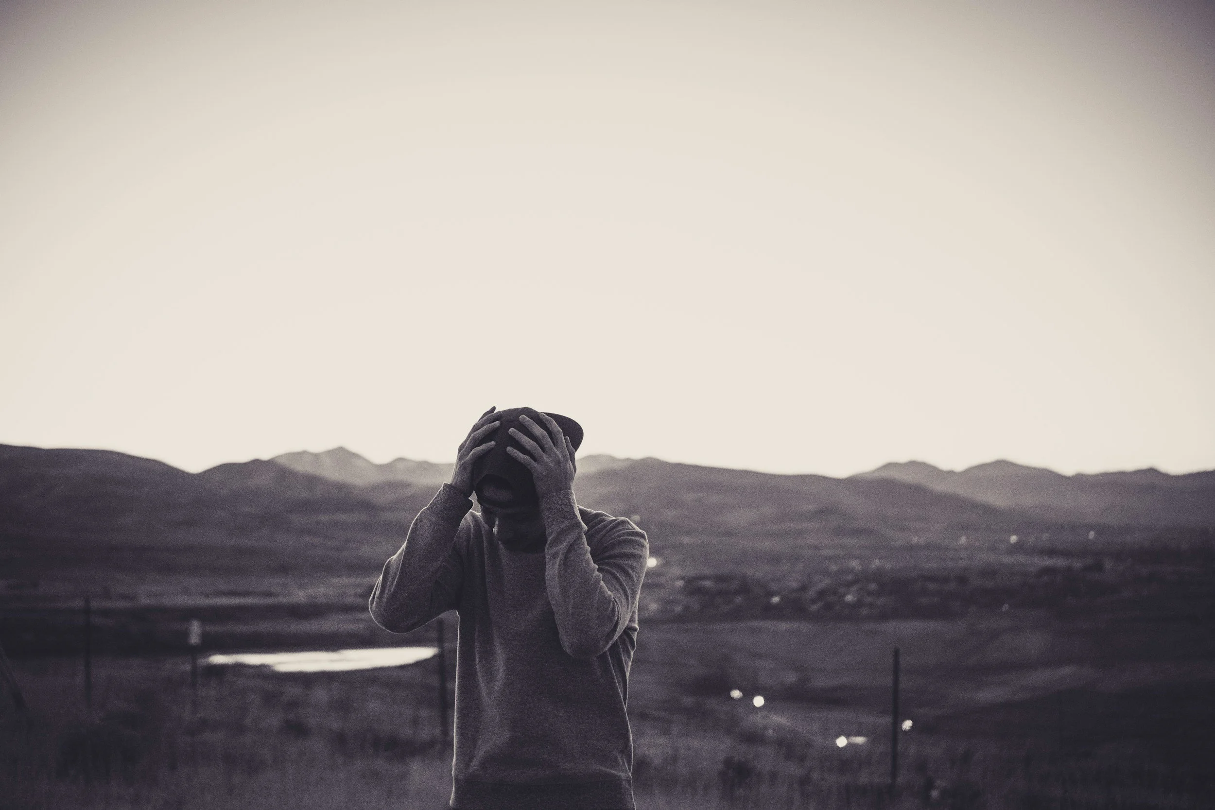 Person holding a hat over their face outdoors with mountains in the background during sunset or sunrise.