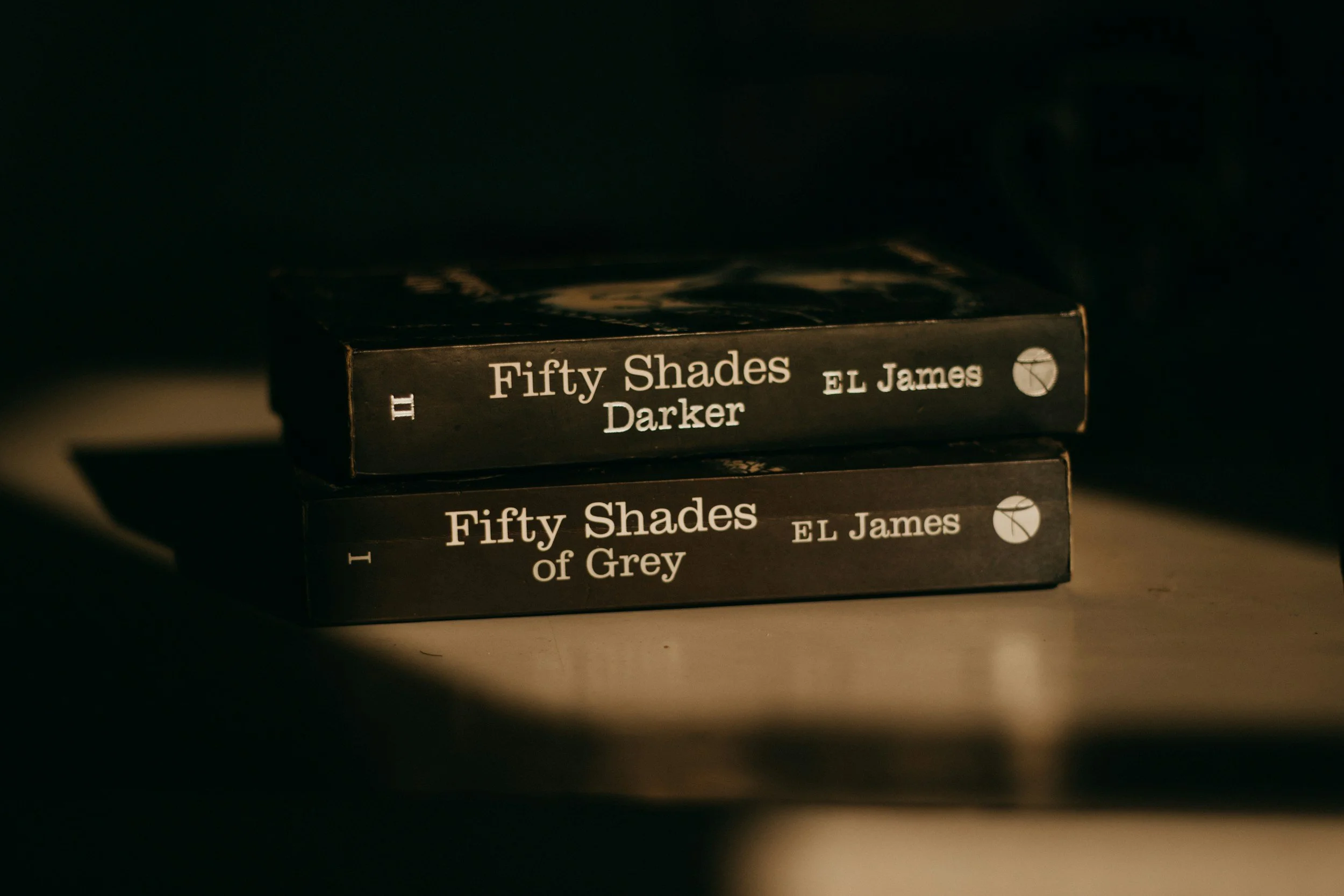 Stack of two black paperback books, titled "Fifty Shades of Grey" by E.L. James, with the second book's subtitle indicating a darker theme.