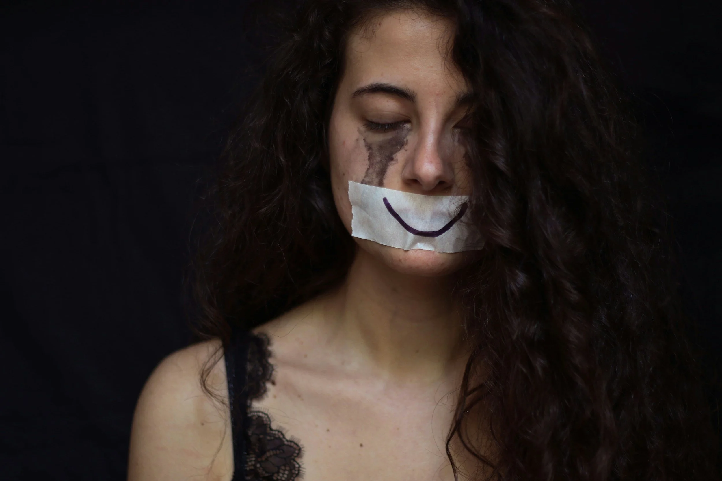 A woman with dark curly hair and light skin, with a painted smile on her lips and a drawn smile on her face mask, eyes closed, against a plain dark background.