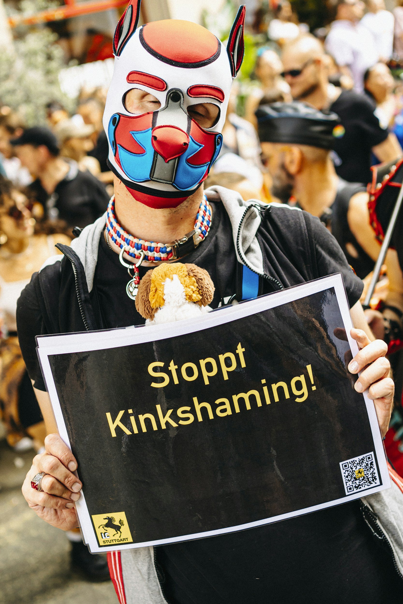 Person wearing a colorful mask, holding a sign that reads 'Stoppt Kuykshaming!'. The person is at a crowded outdoor event, with many people in the background.