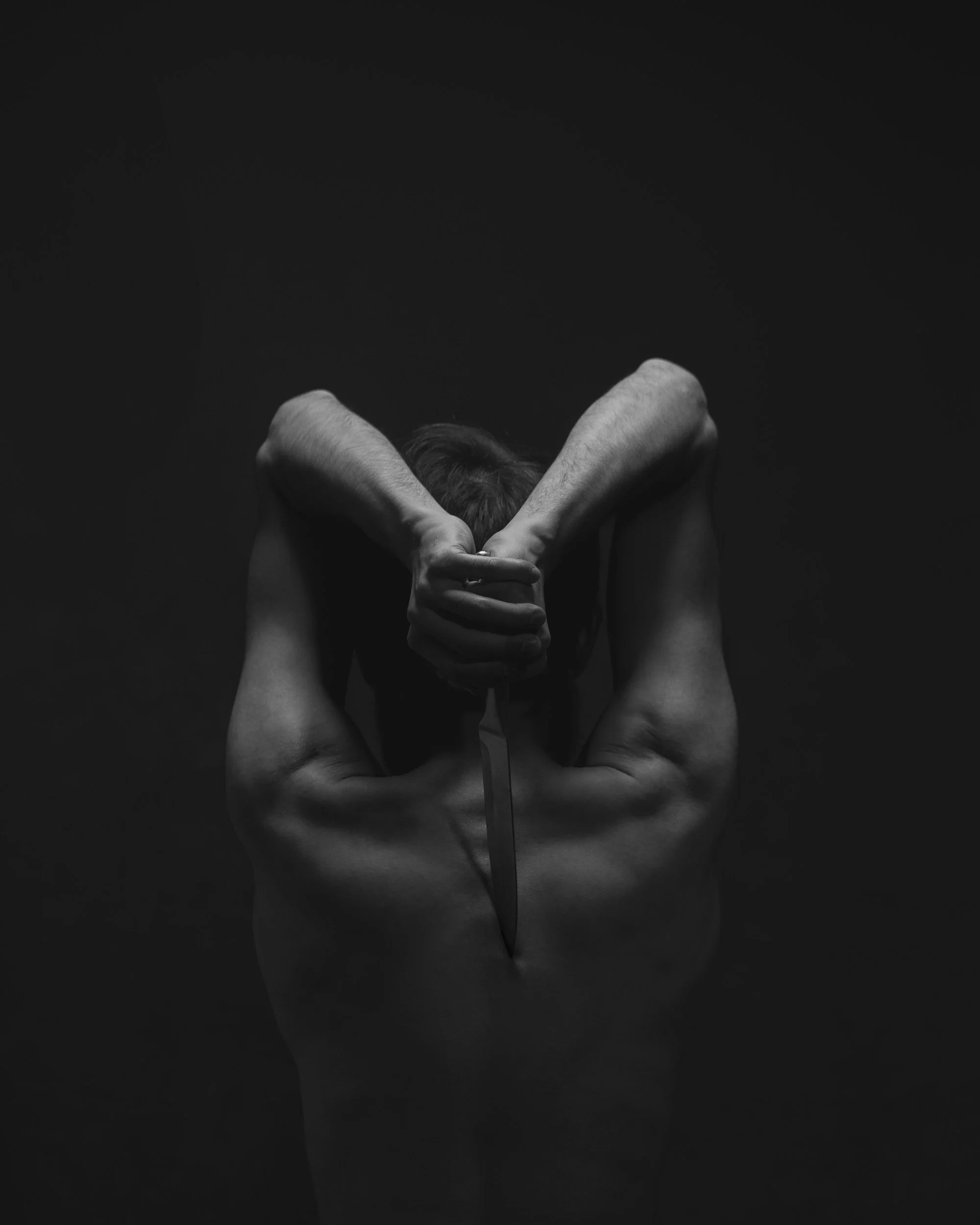 A person with short hair is seated with arms raised, hands clasped behind their head, and holding a knife pointed downward. The person appears to be topless, with a dark background.