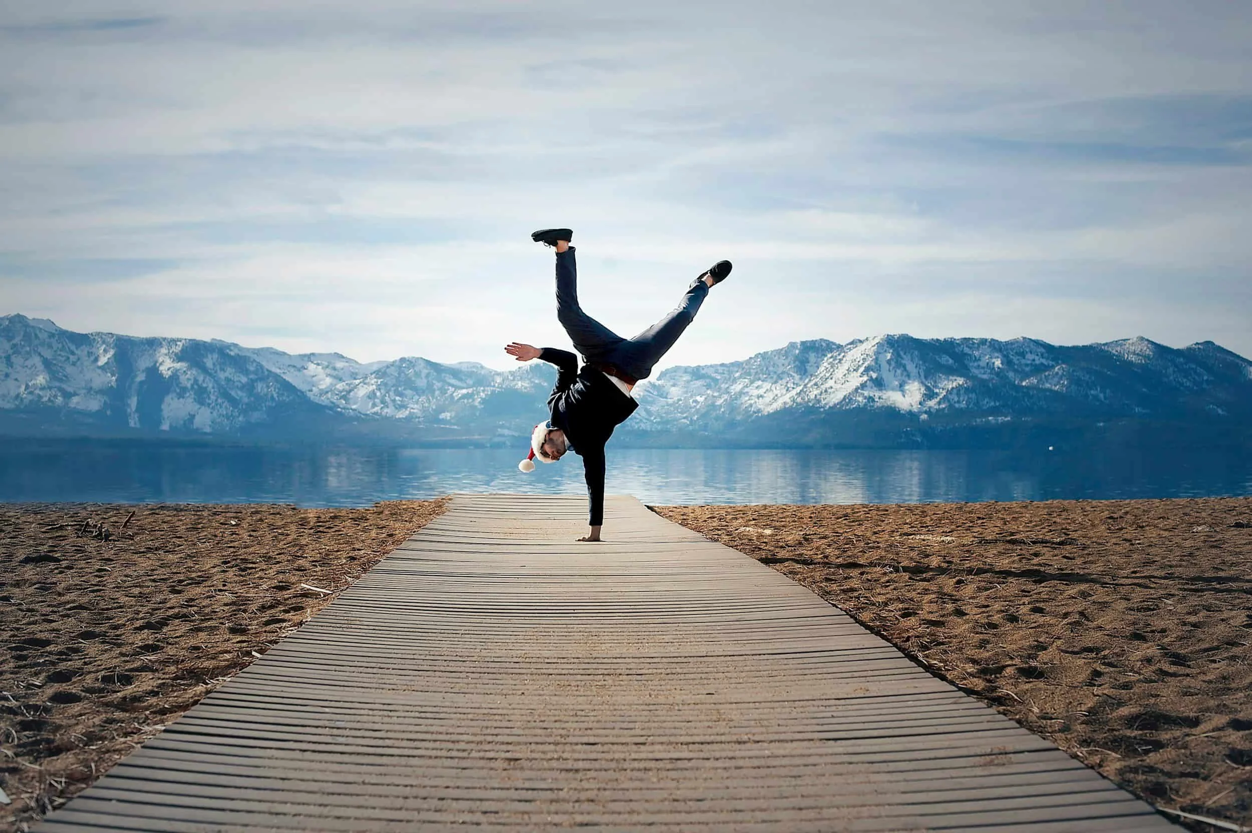 Person in a suit doing a handstand on a wooden path near a body of water with mountains in the background, wearing a Santa hat.