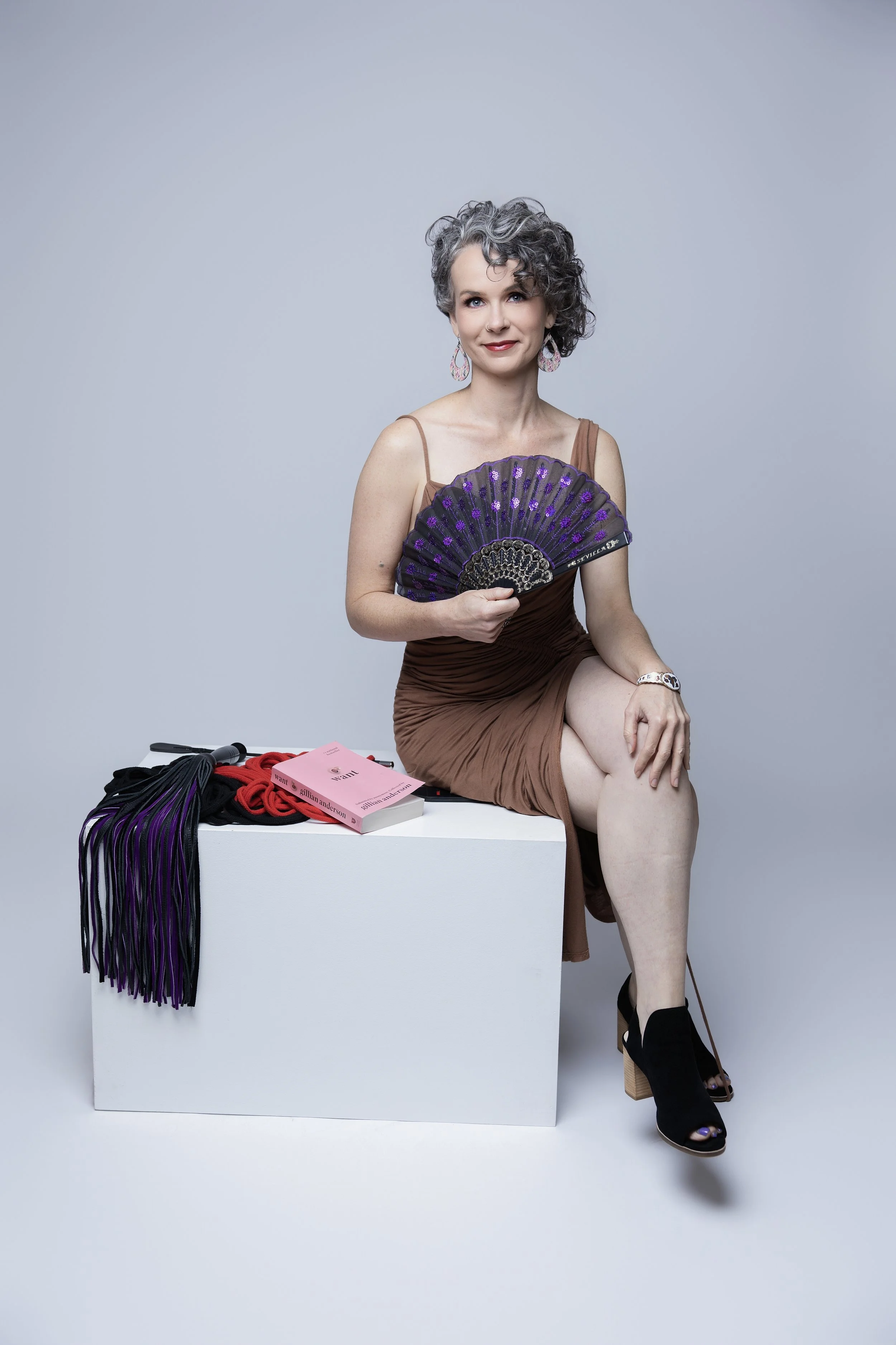 A woman with short gray curly hair holding a purple and black fan, sitting on a white cube with various clothing items and magazines on it, dressed in a brown dress and black high-heeled sandals.