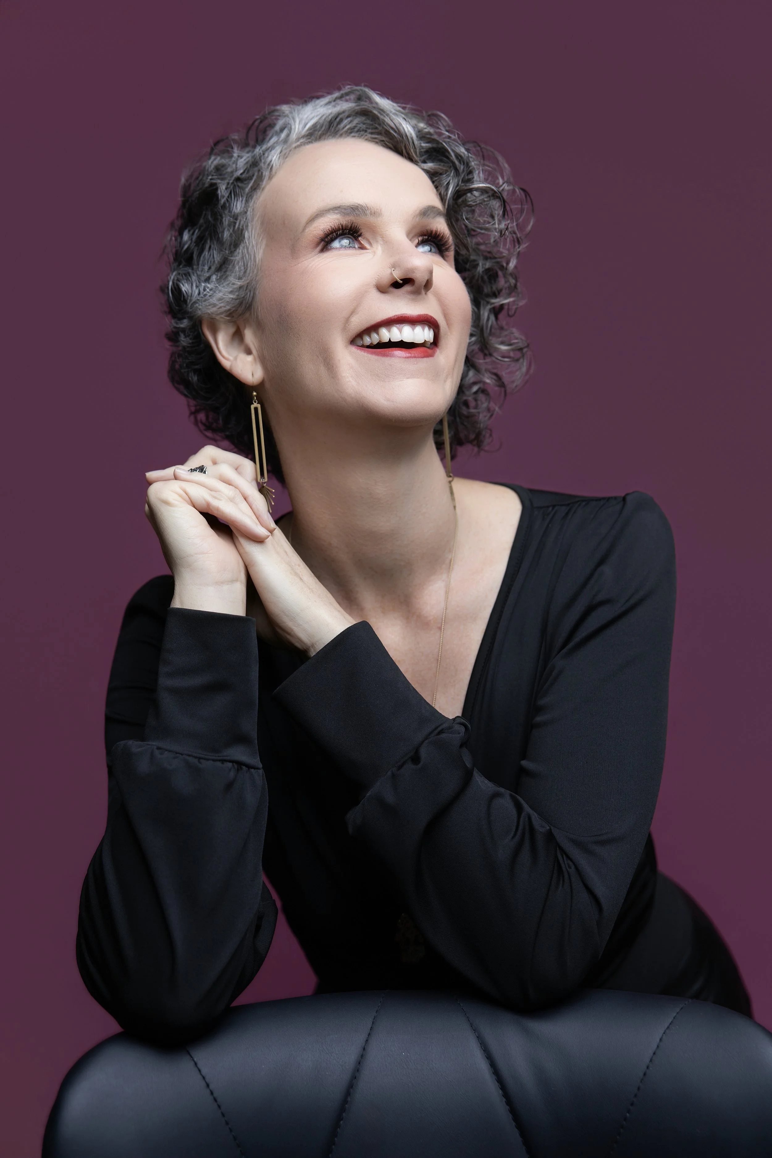 A woman with short curly gray hair, wearing a black long-sleeve top, gold earrings, and a nose ring, smiling and looking up against a purple background.