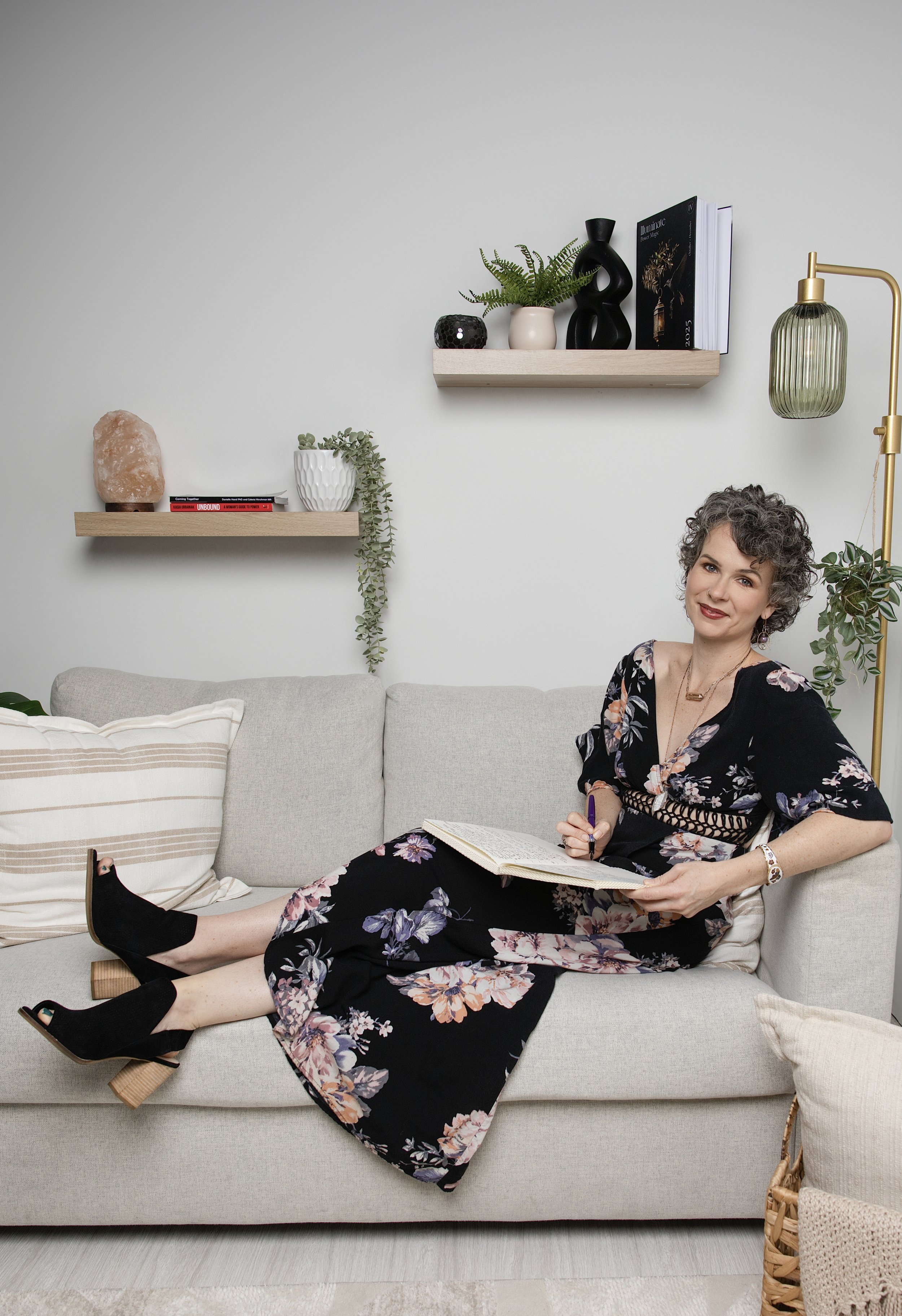 A woman with curly gray hair wearing a black floral dress, sitting on a beige sofa, smiling, holding a notebook and pen, with a sofa cushion and a small pillow visible, in a modern living room with light-colored walls, wooden floating shelves with decorative items, and a gold floor lamp.