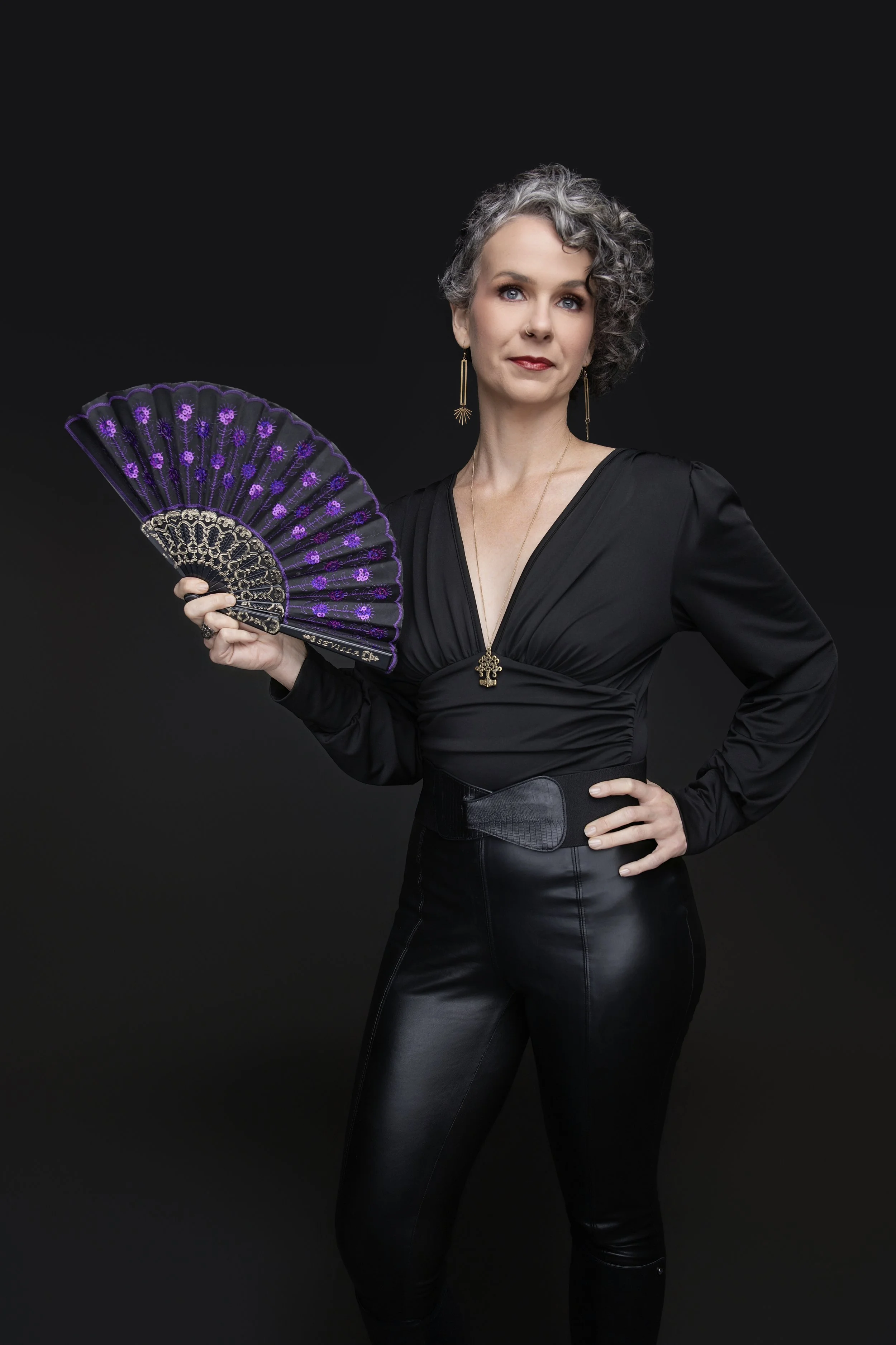A woman with short curly gray hair, wearing a black long-sleeved top and black leather pants, holding a purple and black decorative fan, standing against a solid black background.