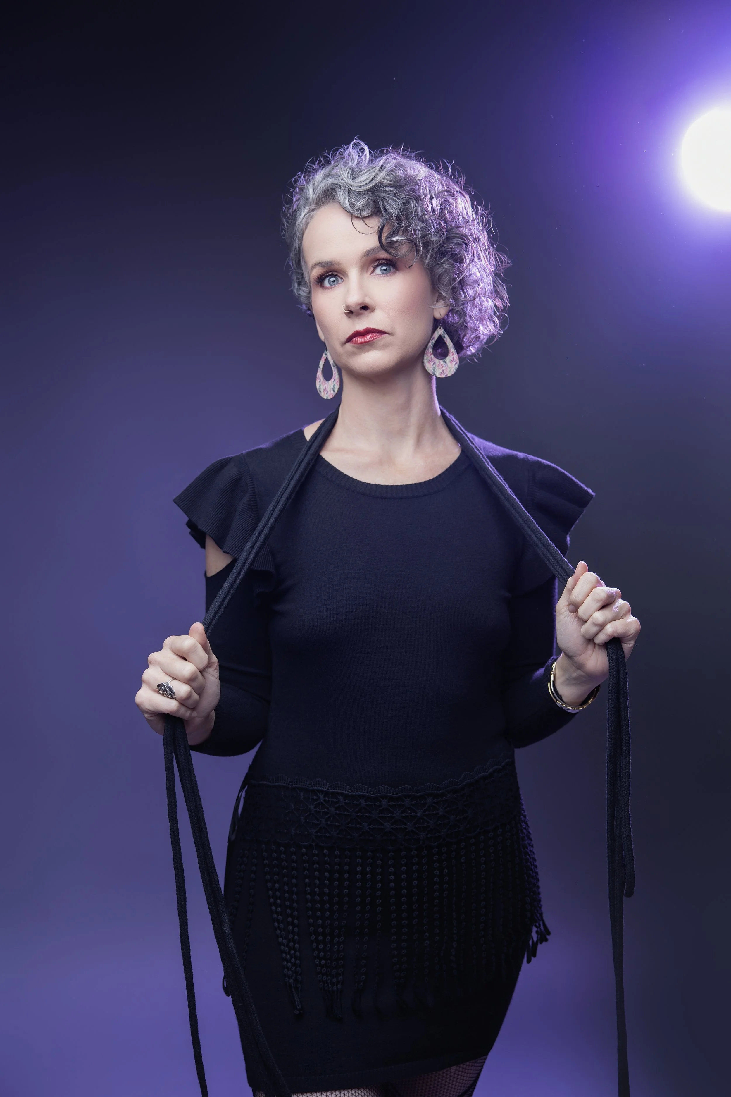 A woman with short, curly gray hair and blue eyes posing against a dark purple background. She is wearing a black outfit with ruffled sleeves, large patterned earrings, and holding a whip in each hand with a serious expression.