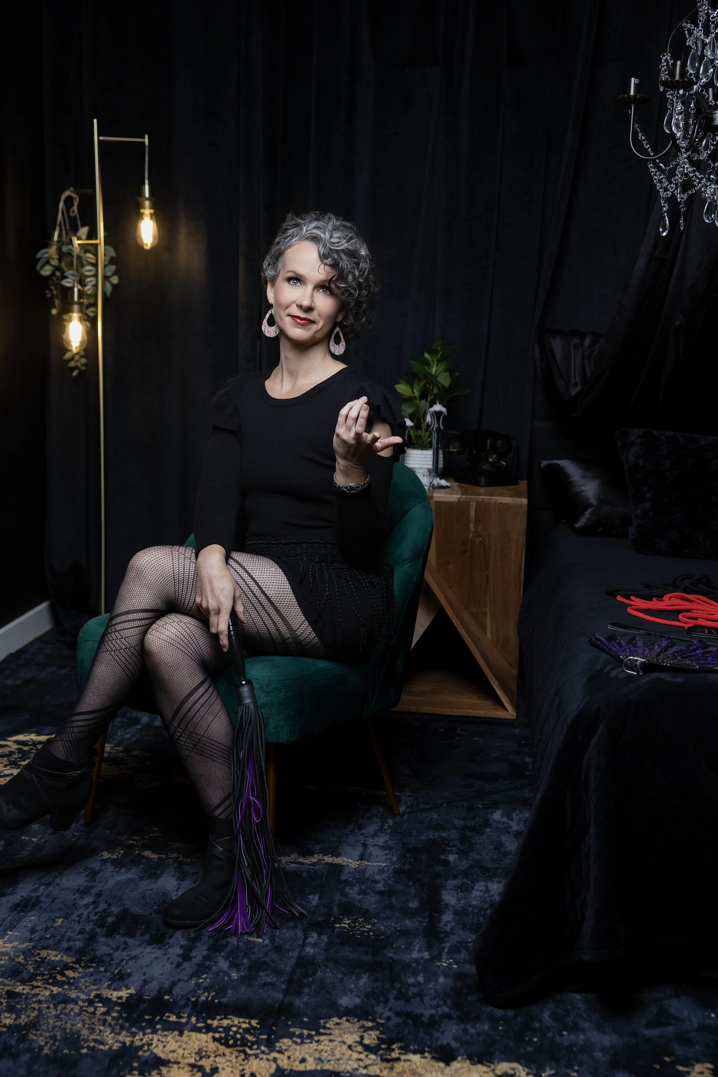 A woman with grey curly hair seated on a green chair holding a whip, wearing fishnet stockings, black boots, and a black top. The room has black curtains, a chandelier, and a side table with a plant. There are BDSM tools on a bed visible on the right.
