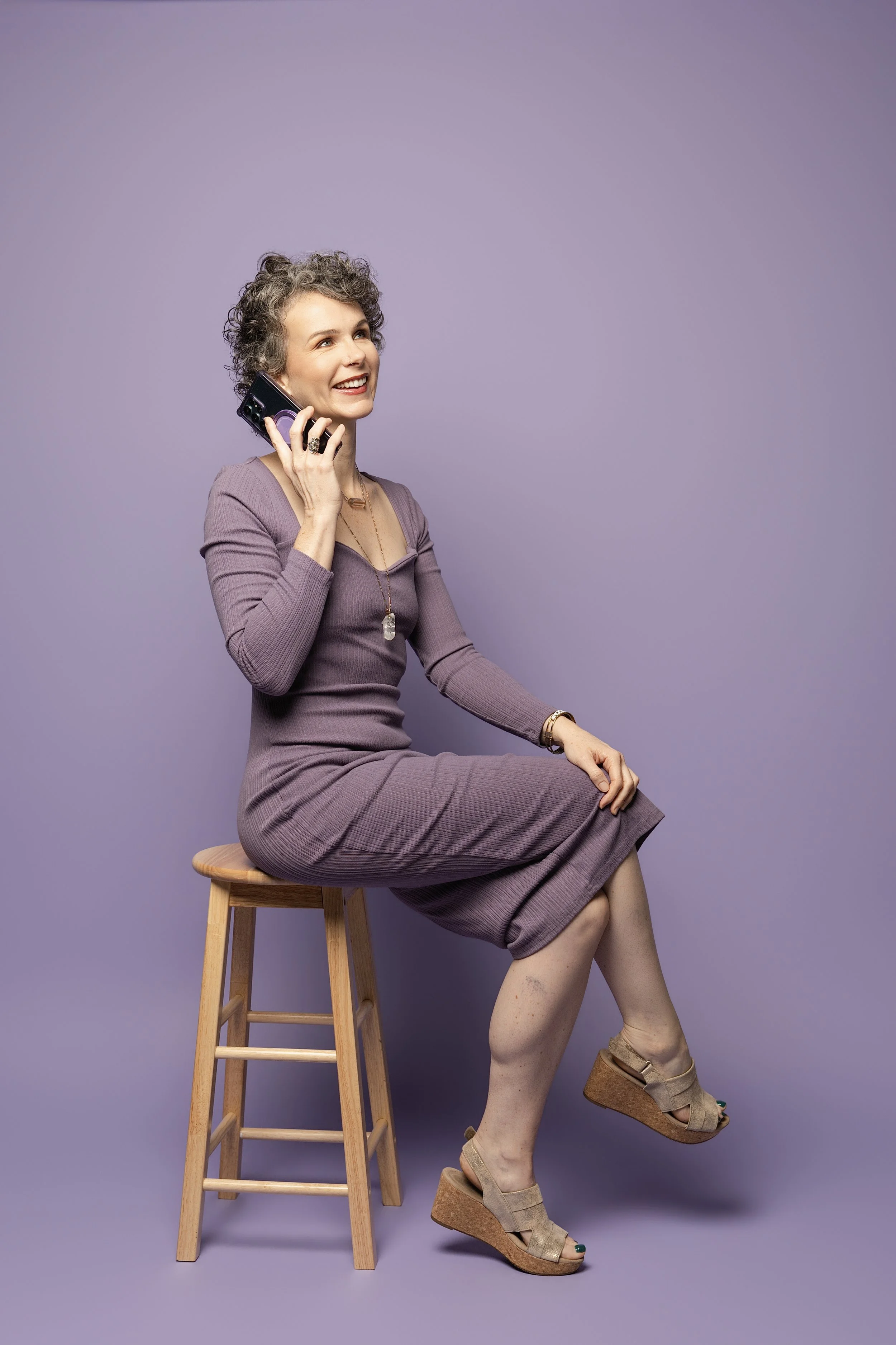 A woman with short curly gray hair, wearing a mauve dress, sitting on a wooden stool with one leg crossed over the other, talking on a phone, against a purple background. She has a cheerful expression and is accessorized with jewelry.