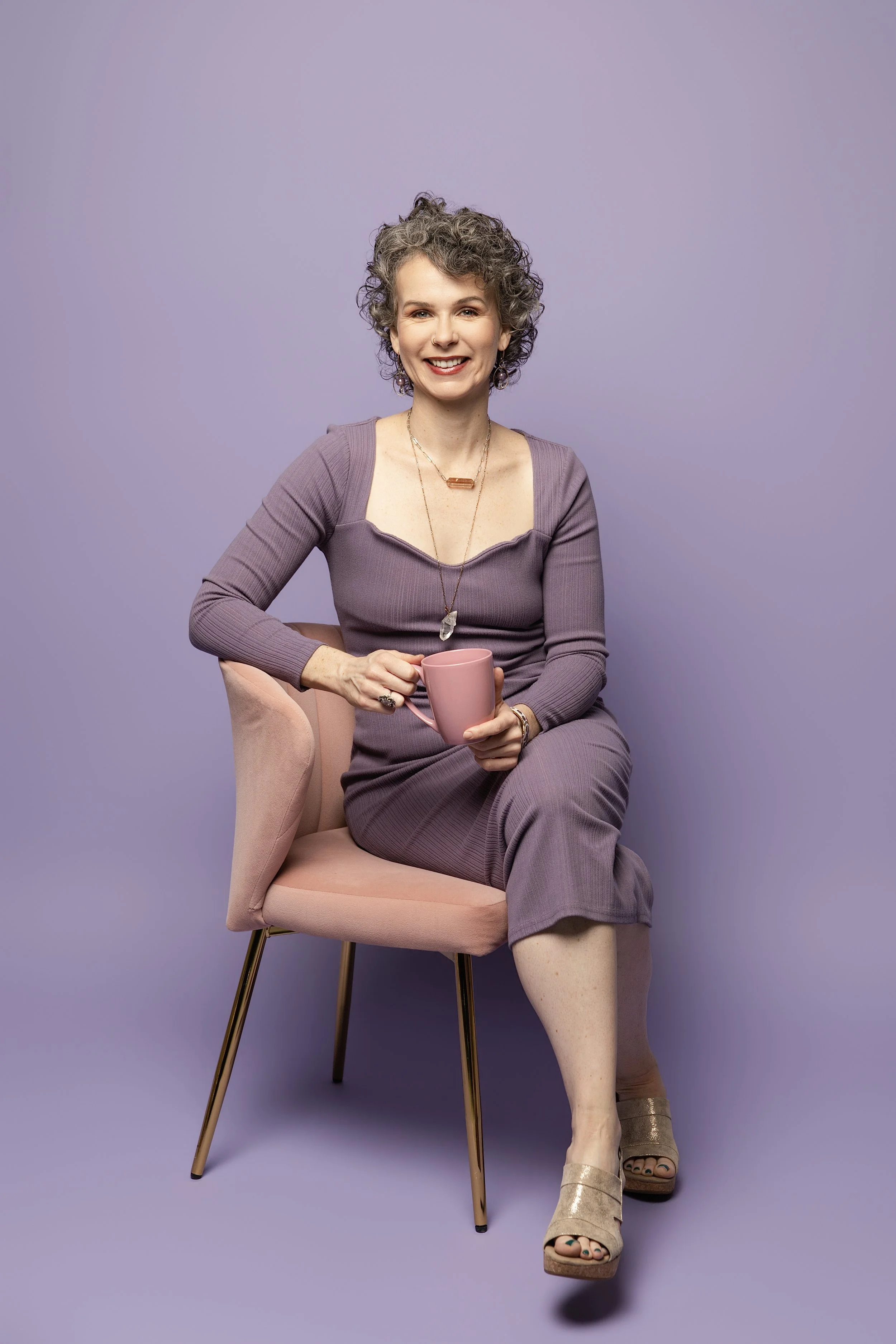 A young woman with short curly dark hair sitting on a pink chair against a purple background. She's smiling, holding a pink mug, and wearing a purple dress, gold sandals, and layered necklaces.