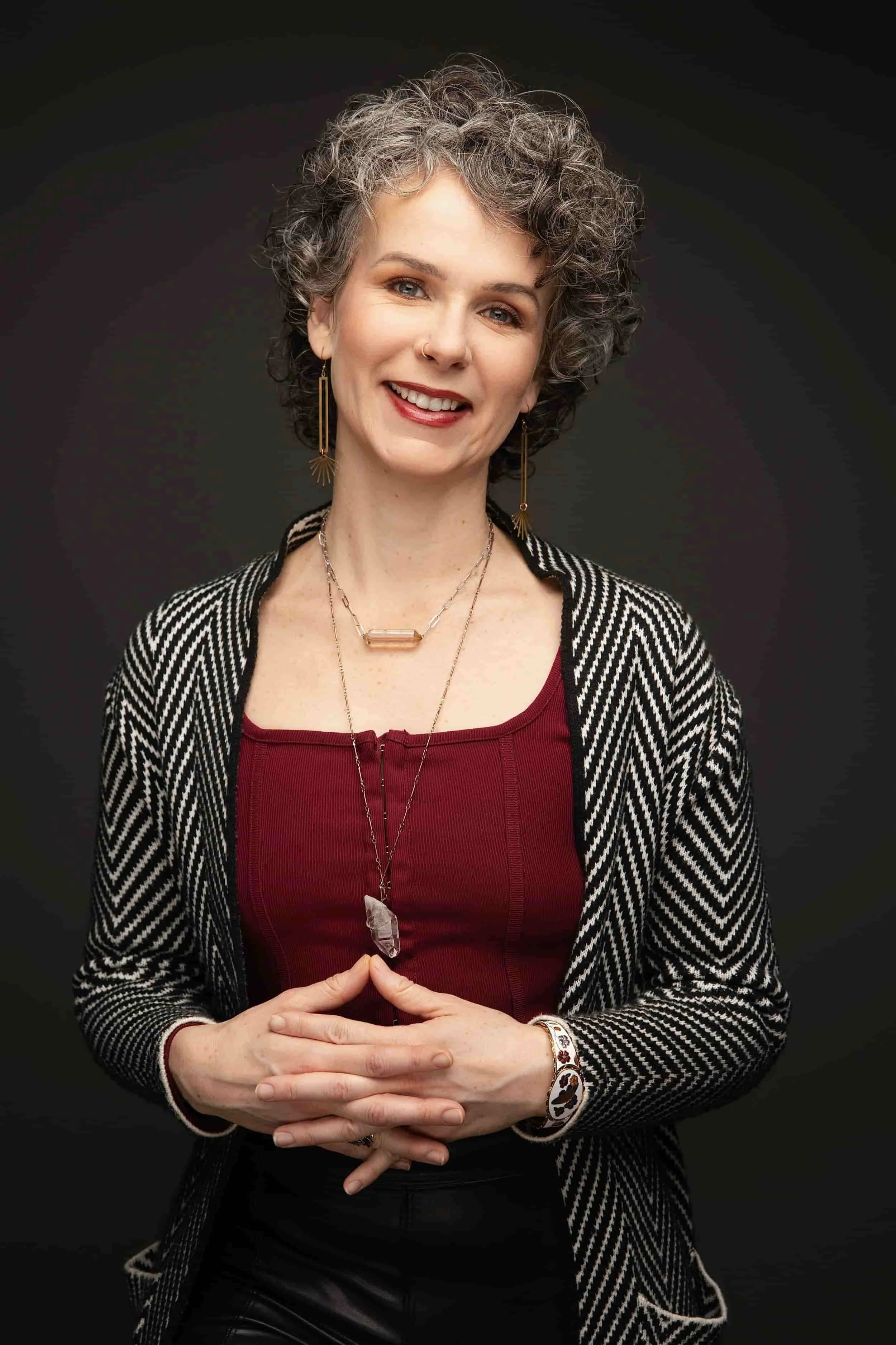 A woman with short, curly gray hair smiling and wearing a black and white striped blazer over a red top with layered crystal necklaces.