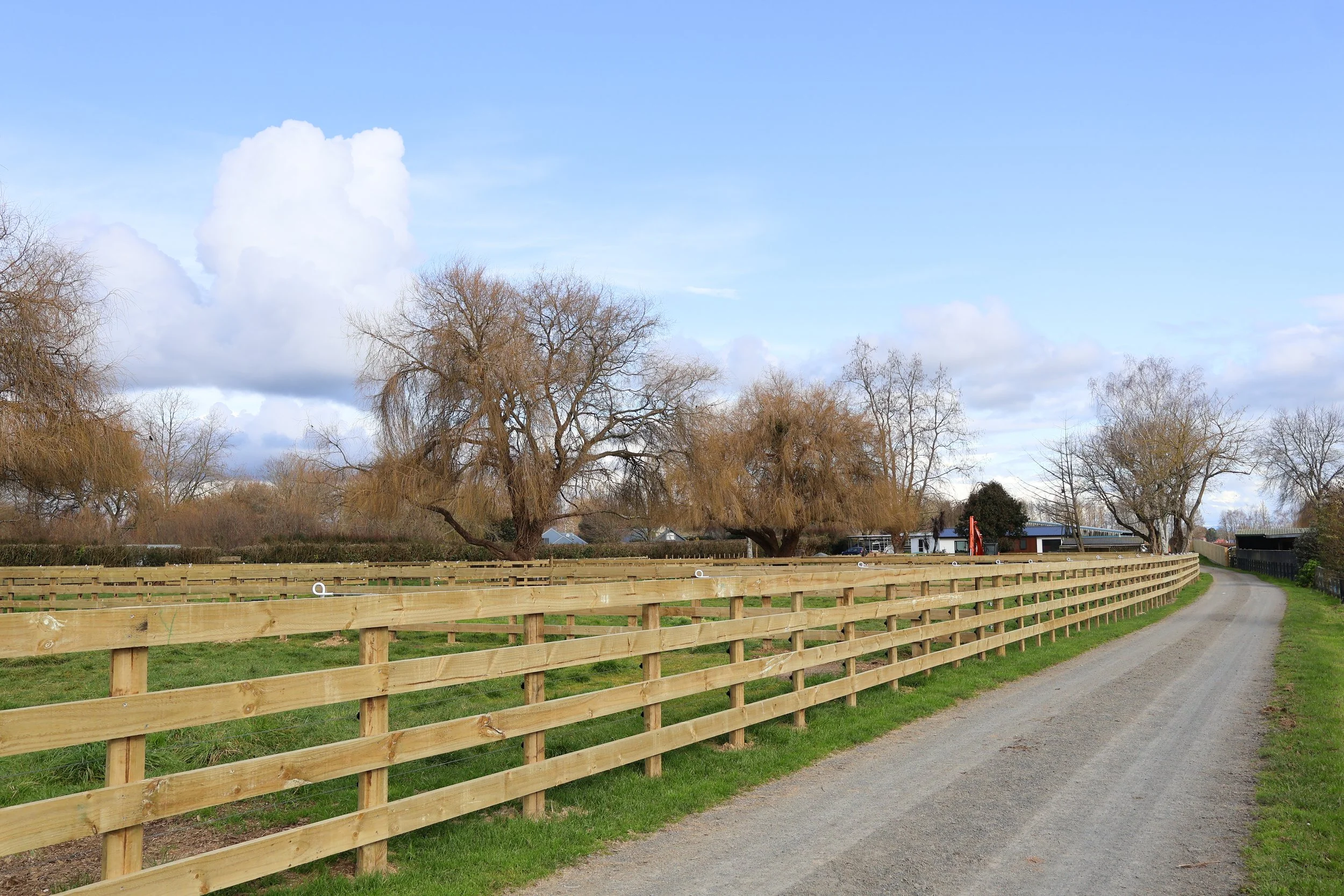 A rural scene with a gravel road curving to the right, a wooden fence running alongside, leafless trees, a blue sky with clouds, and some buildings in the distance.