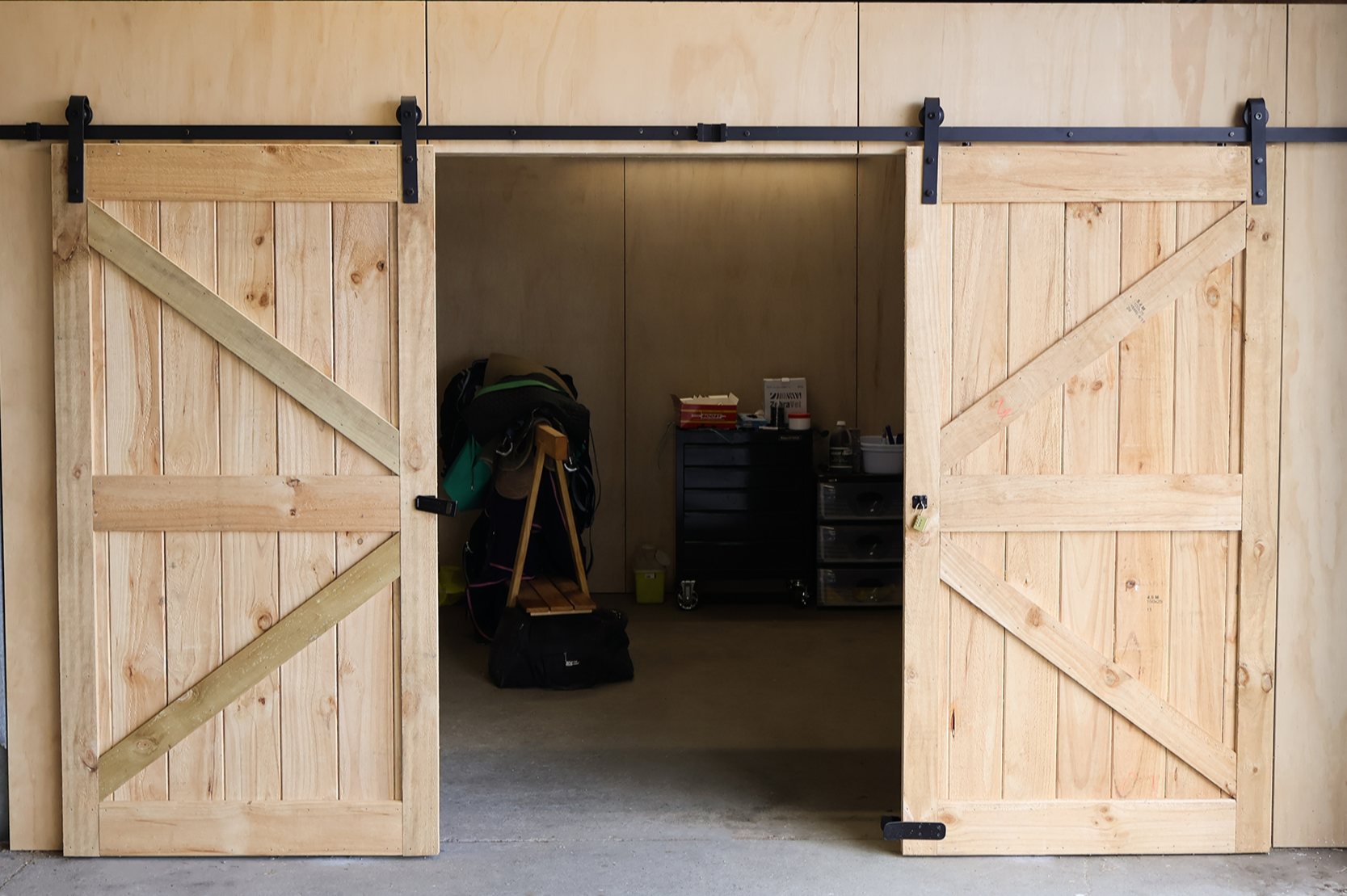 Open wooden barn doors revealing a tack room with a black tool chest, a backpack, and various racing equipment.