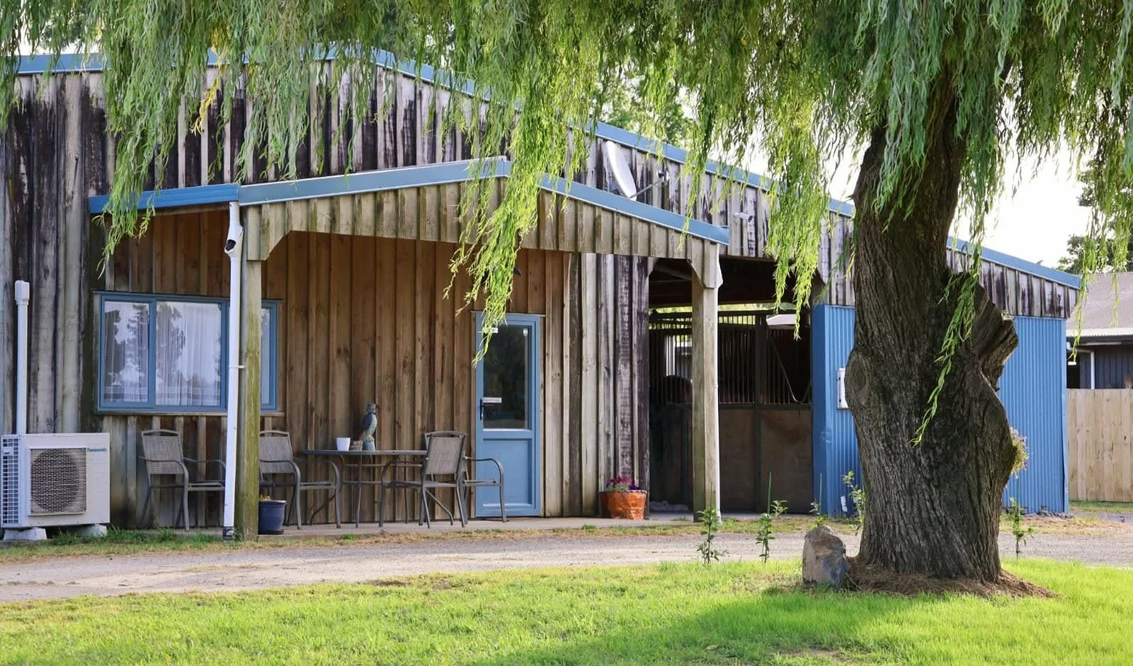 A rustic wooden building with blue accents, surrounded by lush green grass and trees, with outdoor seating and potted plants.