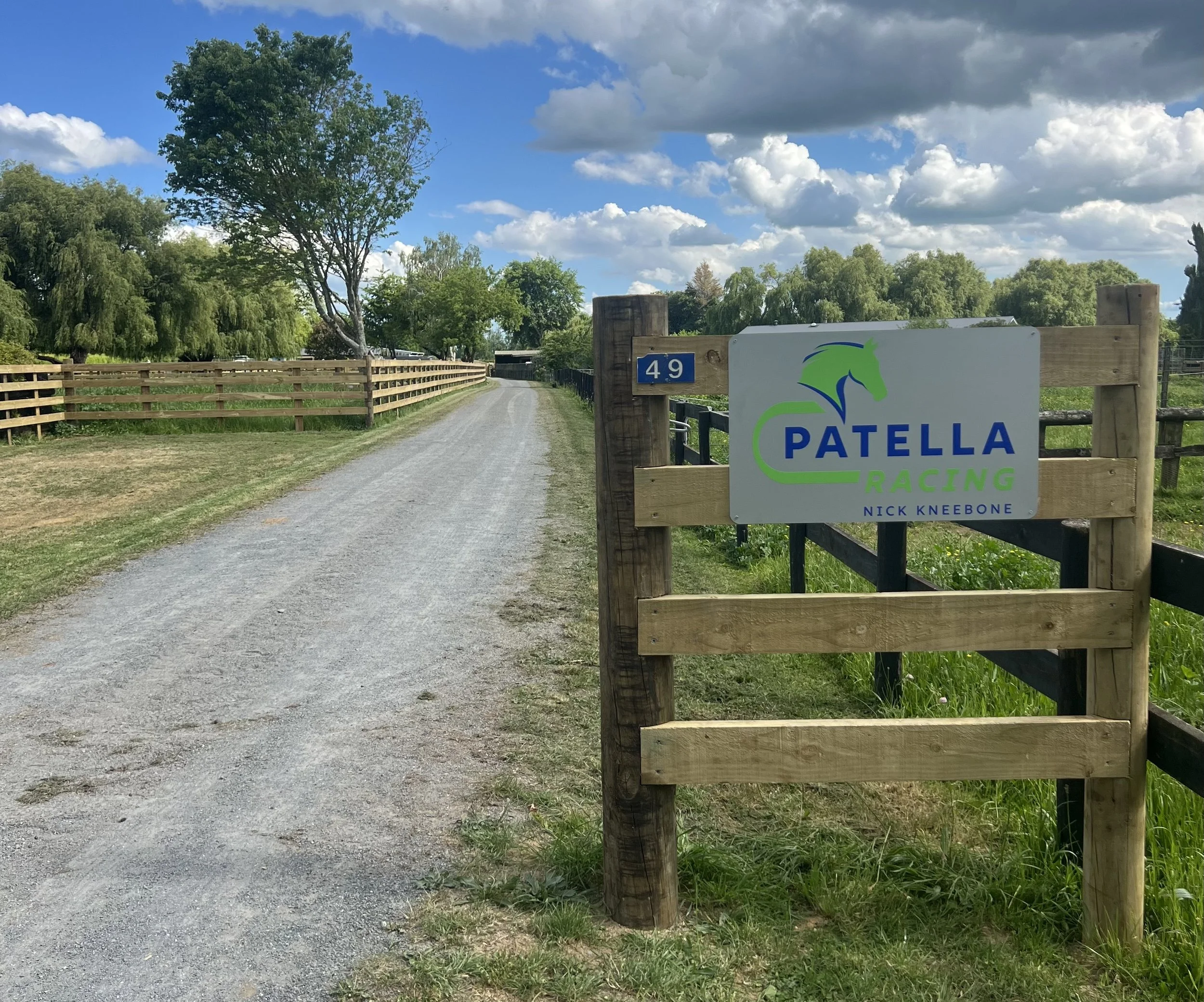 Gravel driveway leading to Patella Racing with a sign that has a green horse logo, the words 'Patella Racing' in blue and green, and the name Nick Knebone, with trees and a blue sky with clouds in the background.