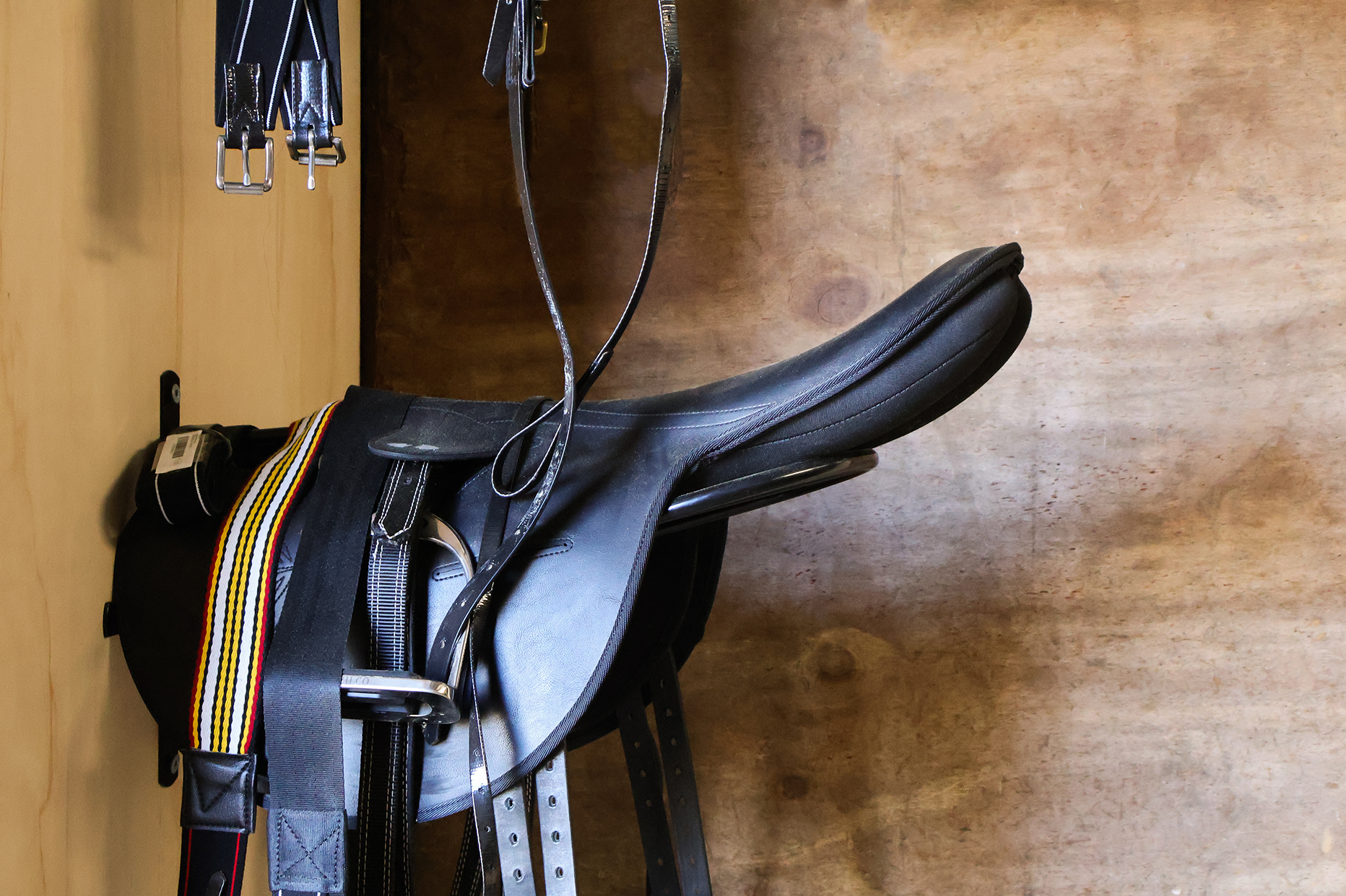 A black horse saddle resting on a saddle stand with straps hanging above, placed on a wooden surface.
