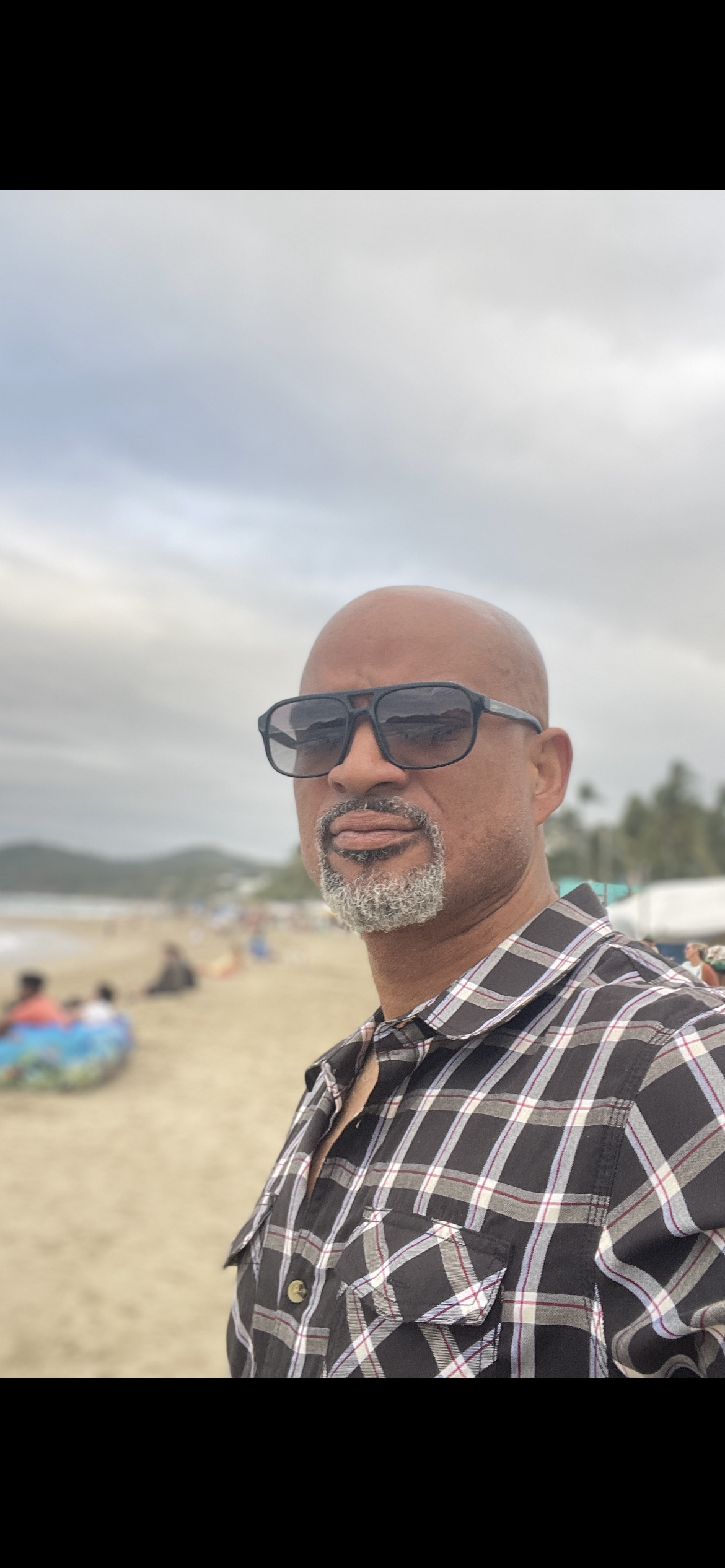 A man with a gray beard wearing sunglasses and a black, white, and red plaid shirt takes a selfie on a beach with cloudy skies and blurred people in the background.