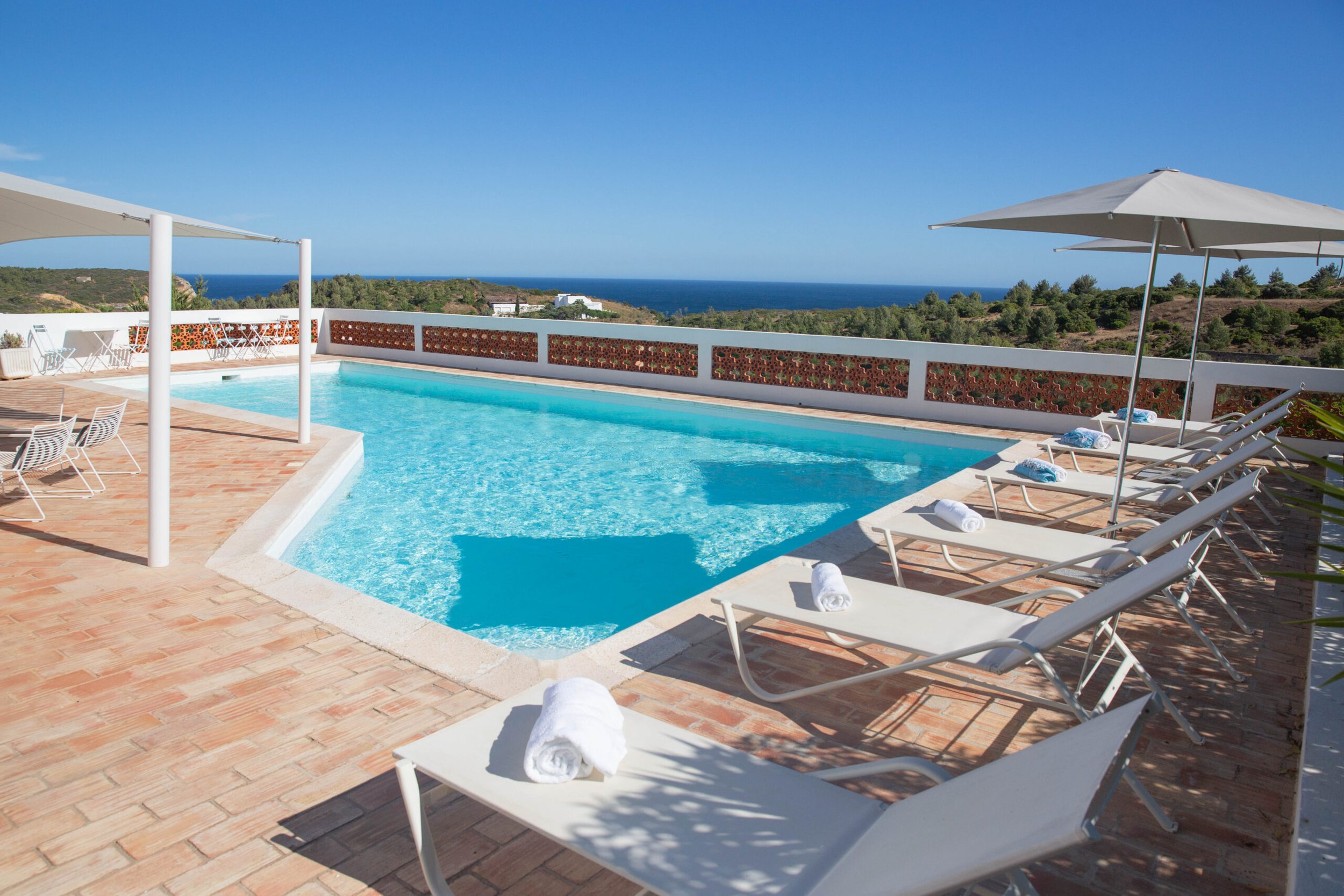 A swimming pool with white lounge chairs and rolled towels around it, overlooking a scenic landscape with green hills, trees, and a distant ocean under a clear blue sky.