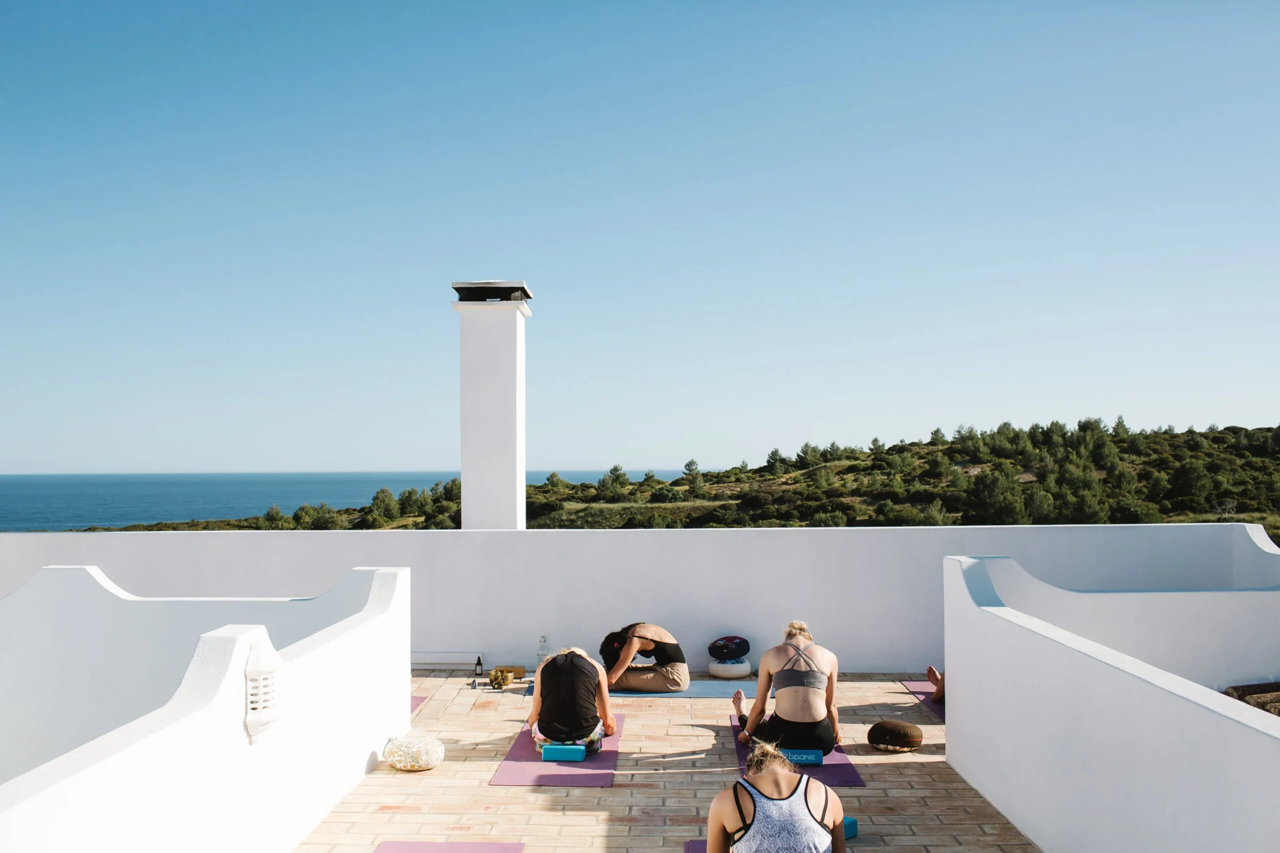 People practicing yoga on yoga mats on a terrace with white walls, overlooking a scenic coastal landscape with green hills and the ocean under a clear blue sky.