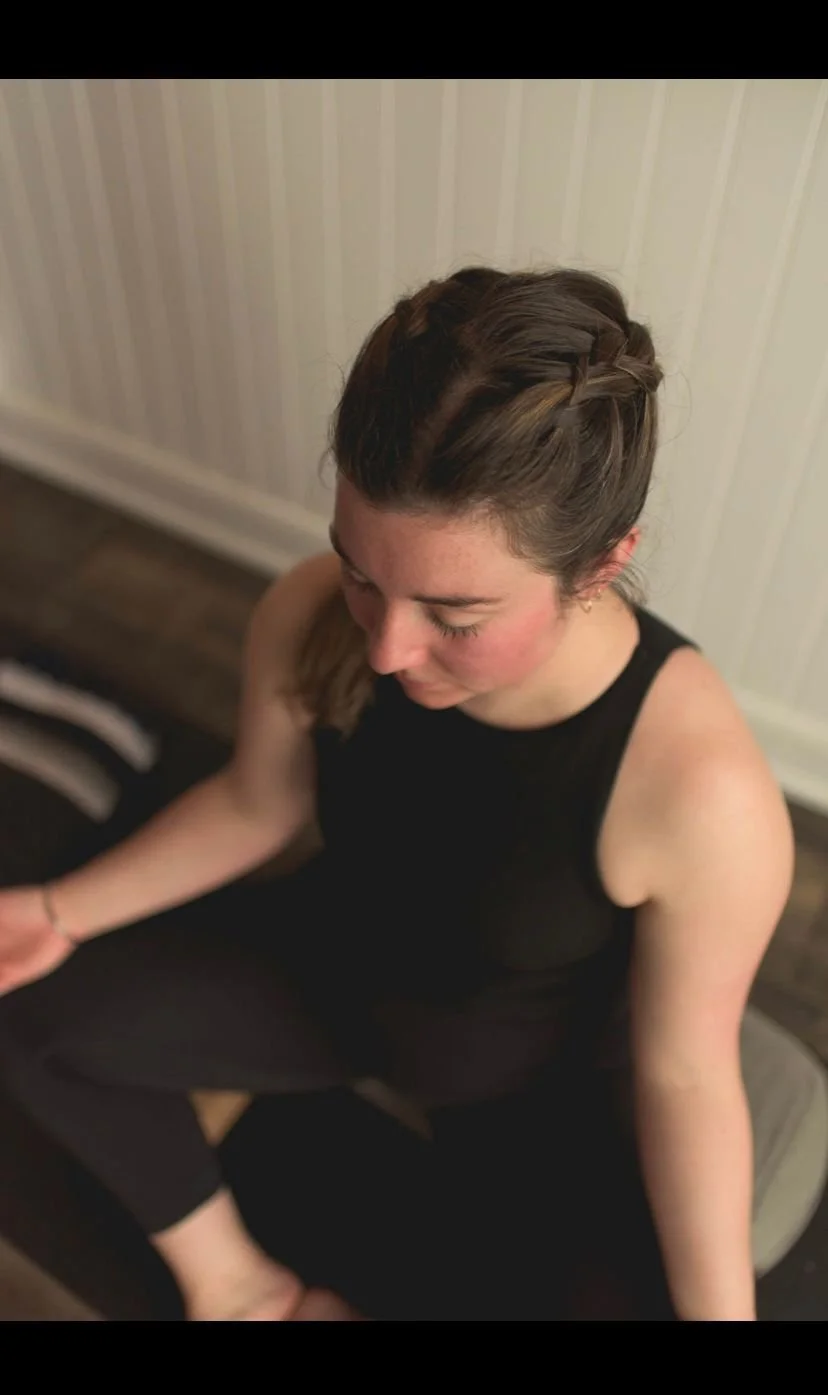 A young woman with brown hair styled in a braid, sitting on a bench, wearing a black sleeveless top and black pants, looking downward with her eyes closed.
