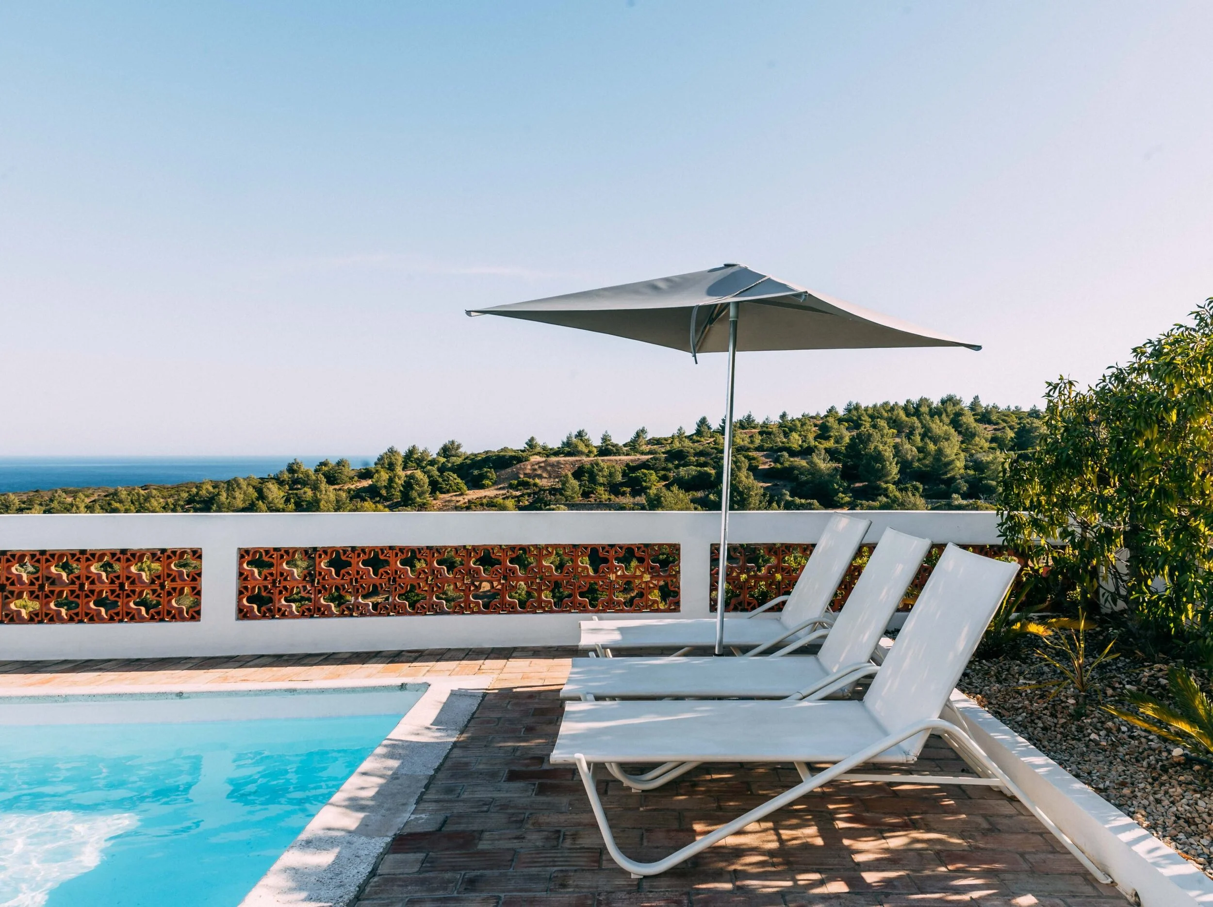 Poolside area with white lounge chairs and a large white patio umbrella on a sunny day, overlooking a hilly landscape with trees and a blue sky.