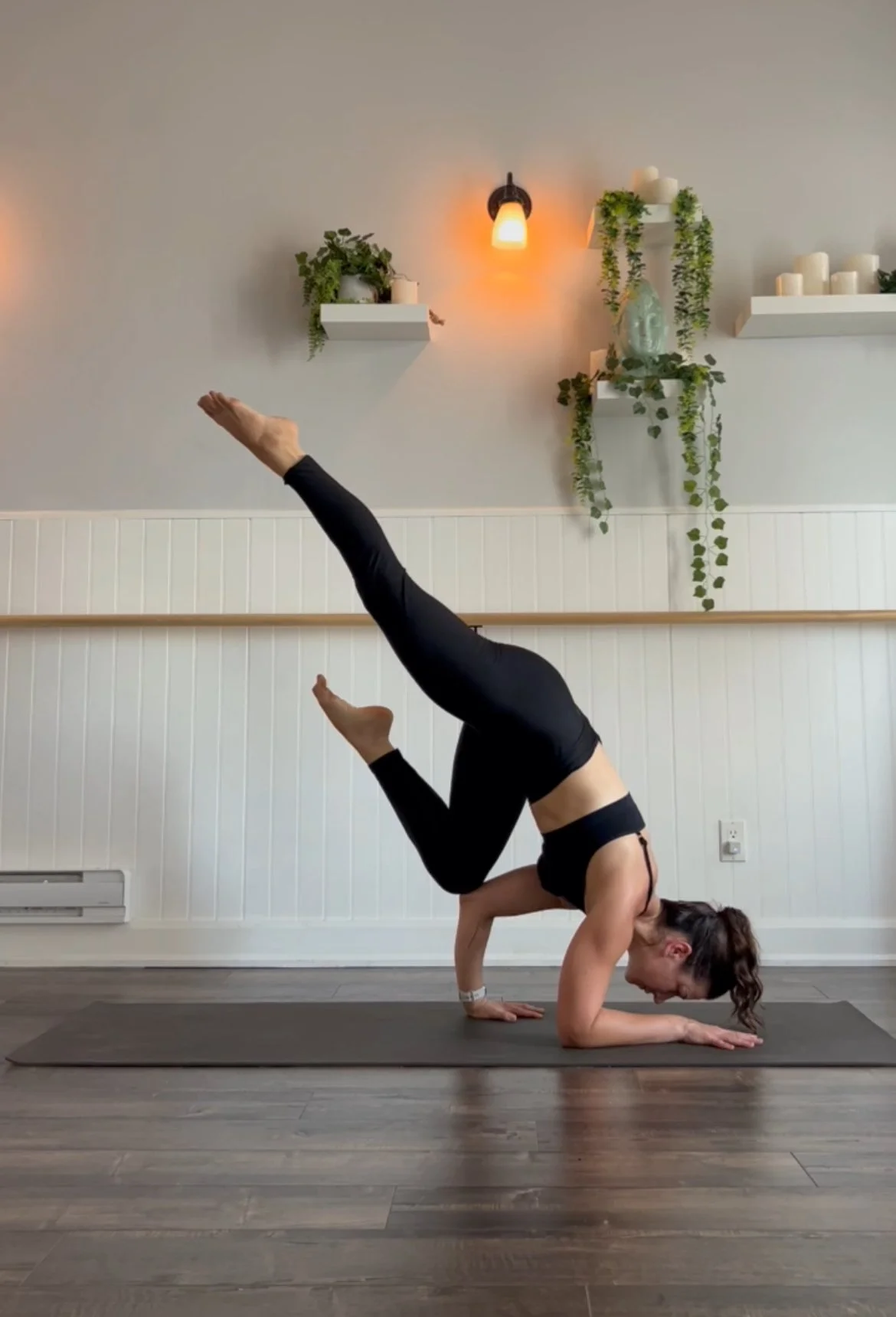 A woman wearing black leggings and a black sports bra practicing yoga indoors in a headstand position on a gray yoga mat. The room has wooden floors, white paneled walls, and decorative shelves with plants and candles on the wall.