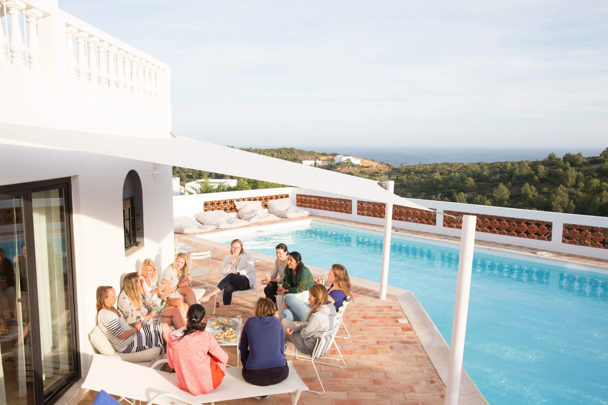 People socializing and sitting around a pool at an outdoor patio with a scenic view of the ocean and green landscape in the background.