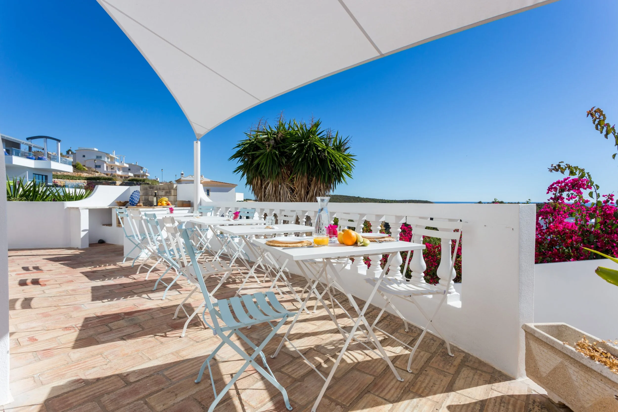 Outdoor patio area with white tables and chairs, set for breakfast with fruit juice, fruit, and bread, surrounded by white walls and colorful flowers, under a large sunshade with a view of the blue sky and ocean in the background.