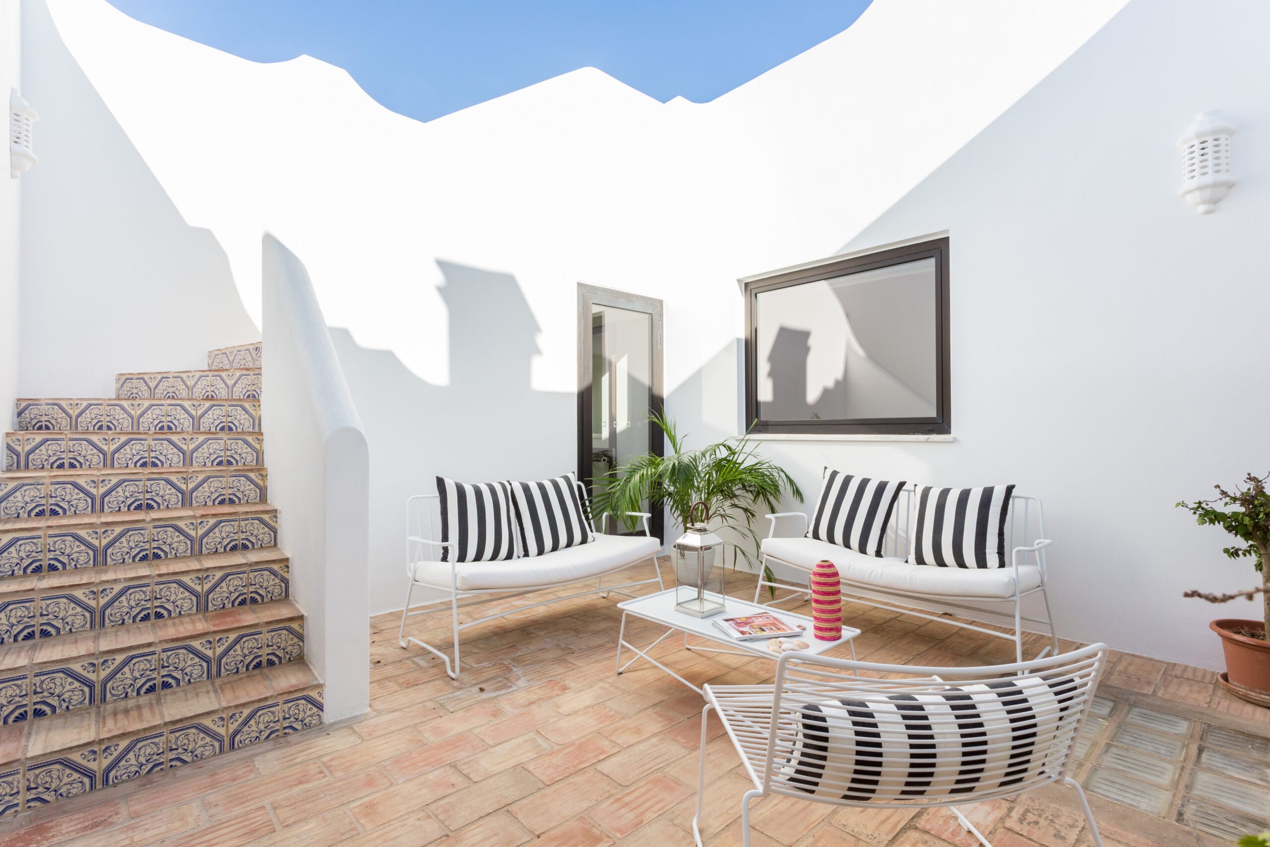 A small outdoor patio area with white walls, two white chairs with black and white striped cushions, a small coffee table with a magazine, a potted plant, and decorative blue and white tiled stairs leading up on the left. There are two large windows 