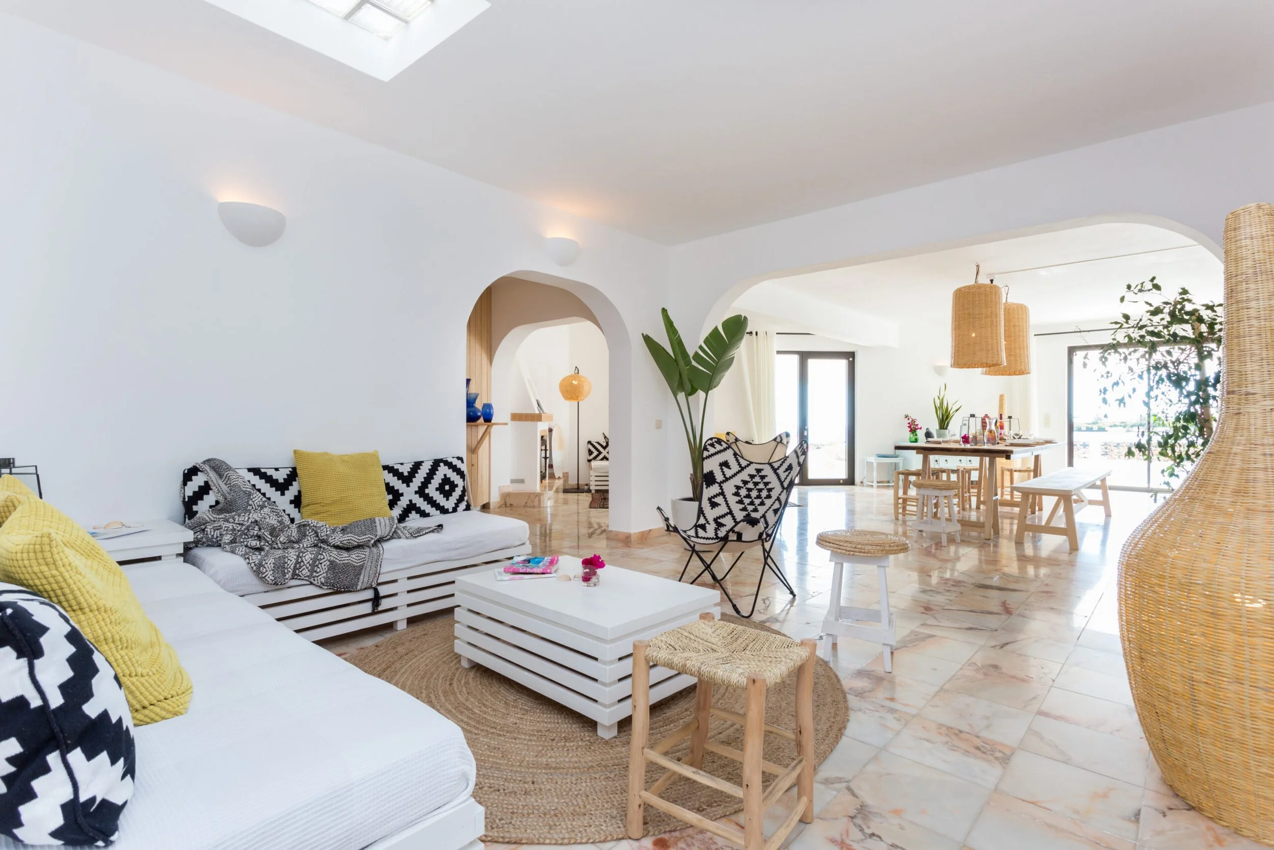 Bright and airy living and dining area with white walls, large windows, and rattan accents. Includes a white sofa with yellow and black decorative pillows, a coffee table, woven pendant lights, and a large plant.