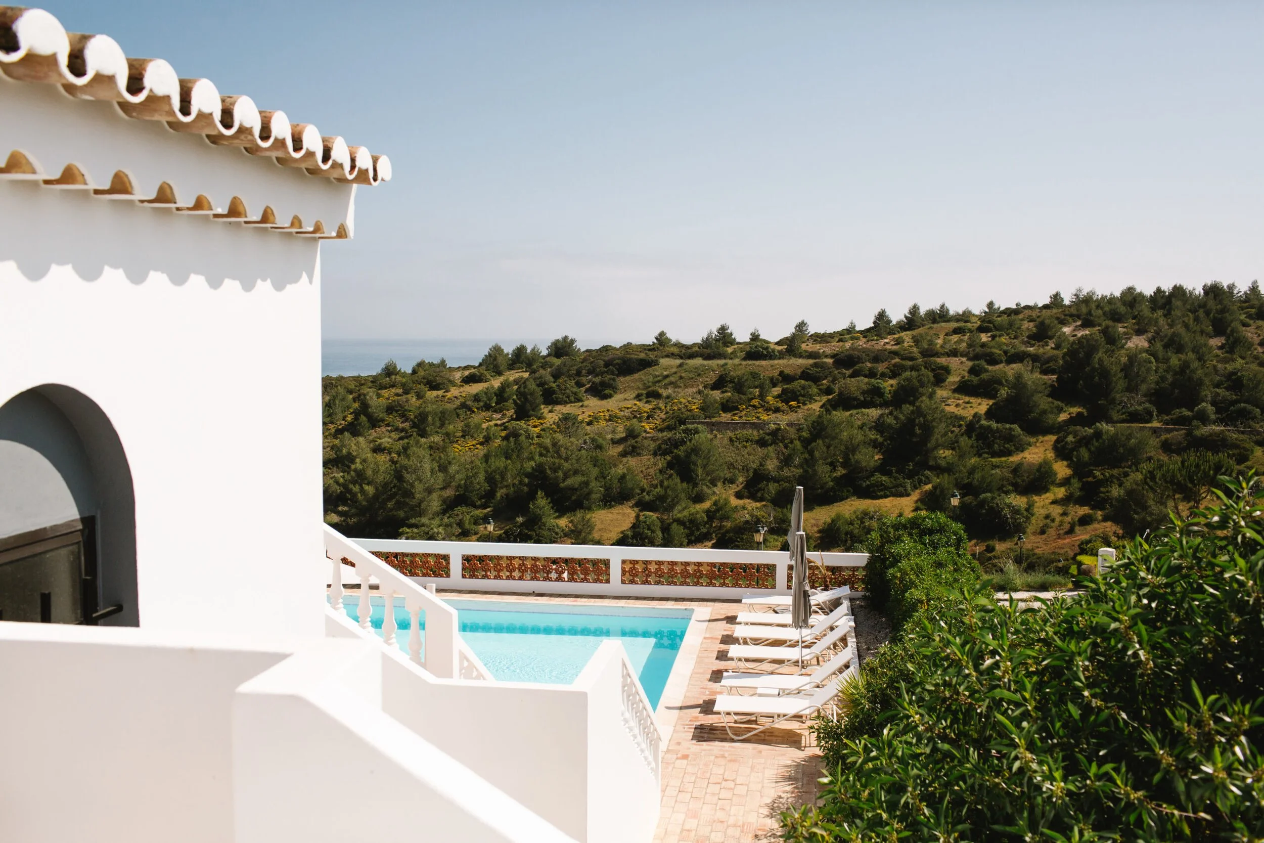 View of a white building with a terracotta-tiled roof, a swimming pool, poolside lounge chairs, greenery, and a scenic hillside landscape under a clear sky.