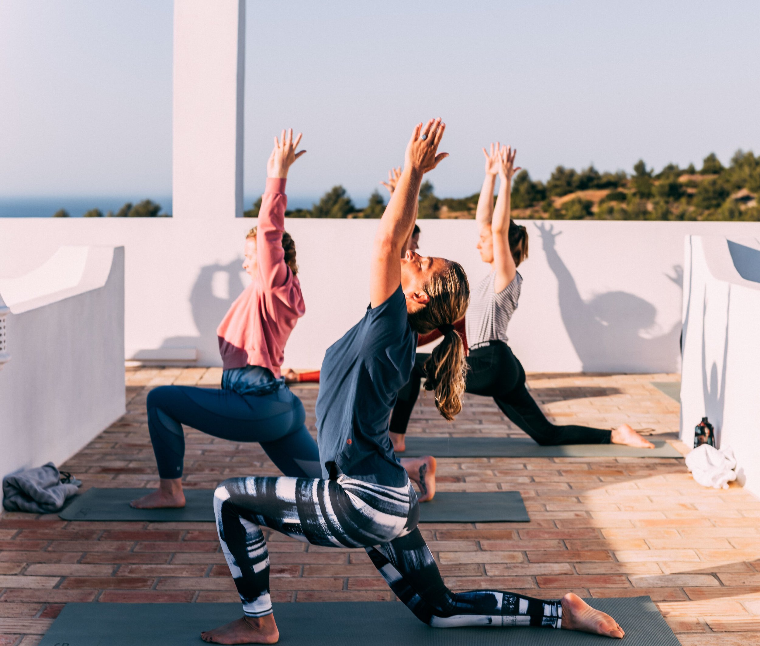 Group of people practicing yoga outdoors on a rooftop, performing a yoga pose with hands raised and one knee on the ground, with a scenic view of trees and hills in the background.