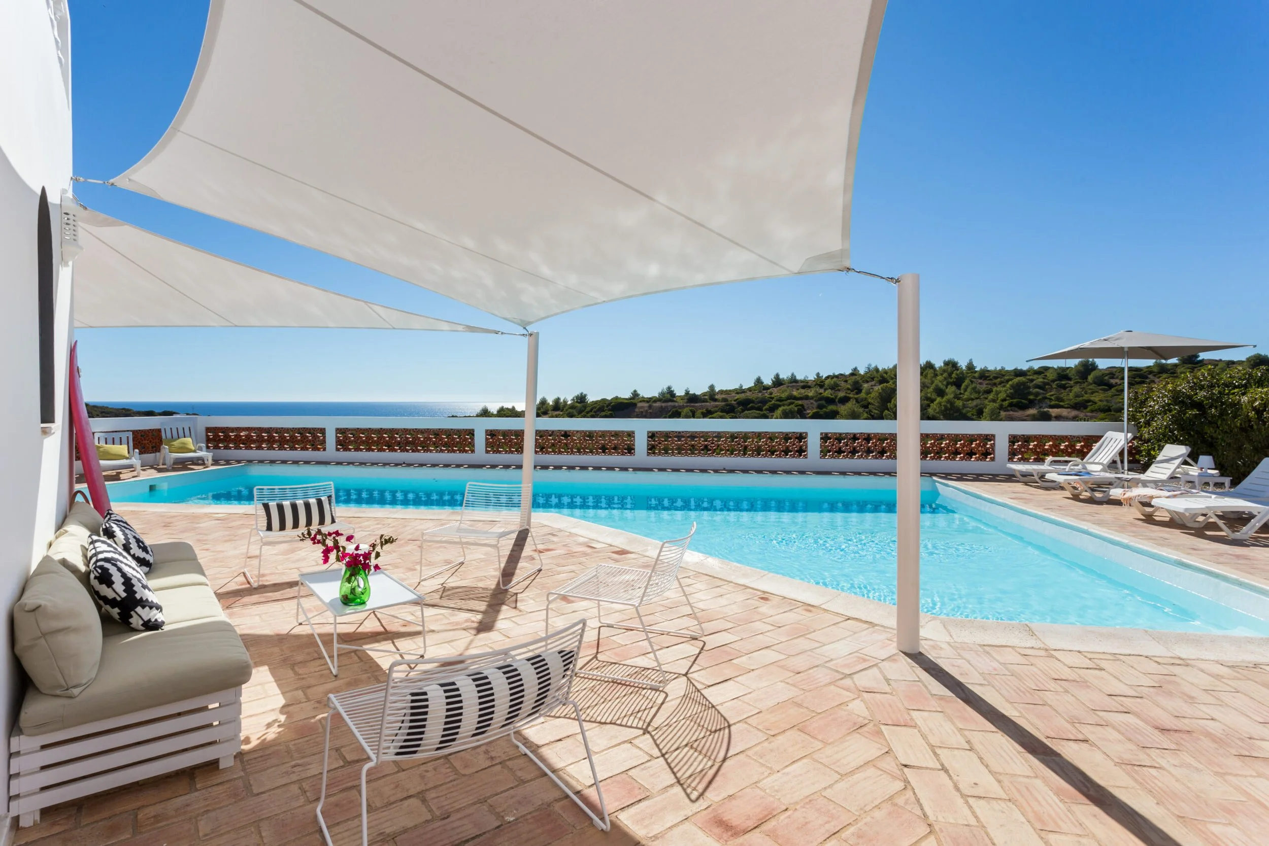 A backyard pool area with white lounge chairs, a sofa with black and white pillows, small tables with a green vase and pink flowers, and large white umbrellas providing shade. The area overlooks green hills and blue ocean in the distance, under a cle