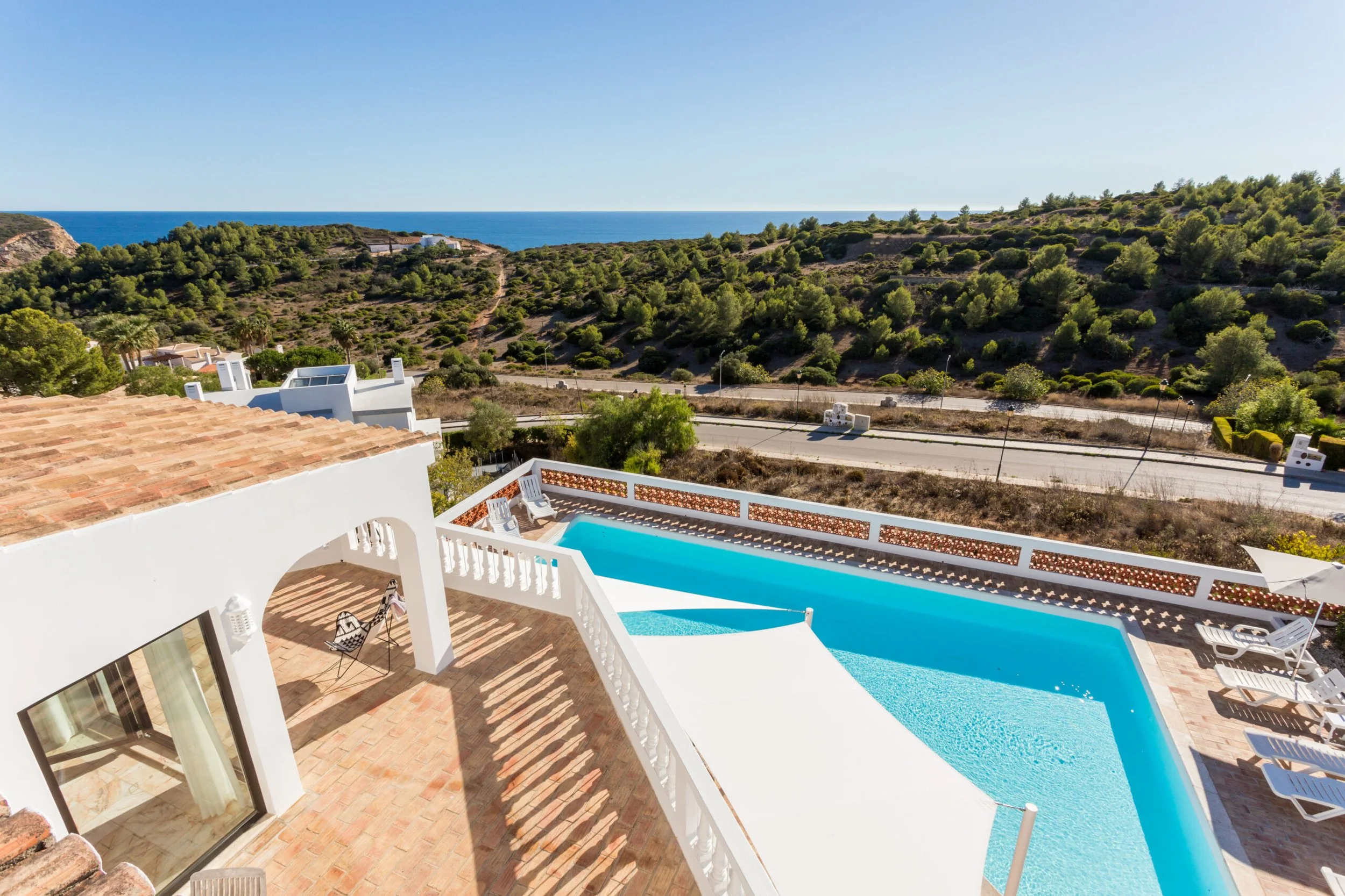 View from a balcony showing a backyard with a swimming pool, tiled patio, and outdoor seating area. Beyond the yard, there is a winding road, a hillside with lush green trees, and the ocean in the distance under a clear blue sky.