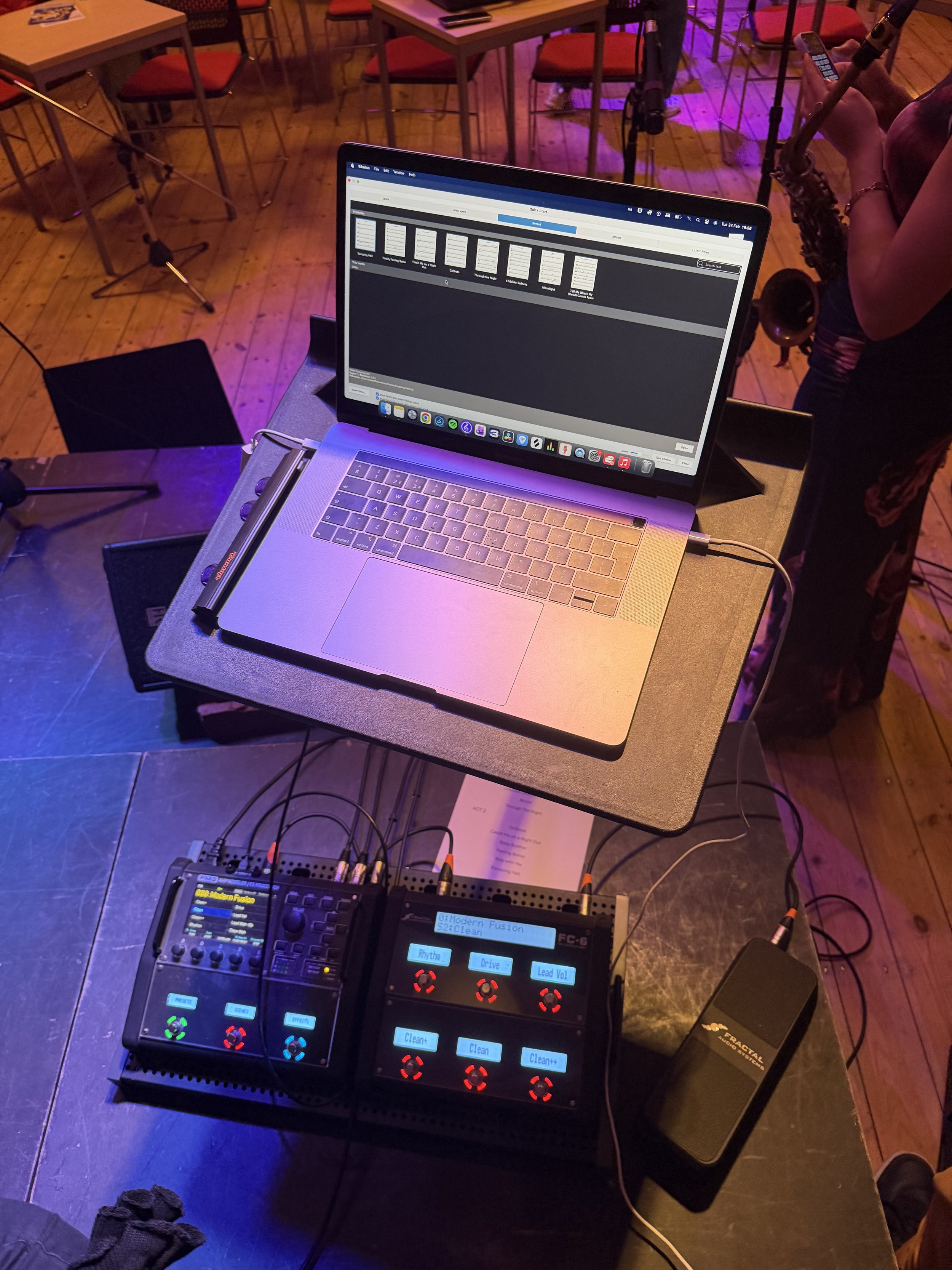 A music setup with a laptop on a black table, a digital audio mixer with colorful buttons nearby, and a person holding a saxophone, at a venue with chairs and tables, and purple lighting.