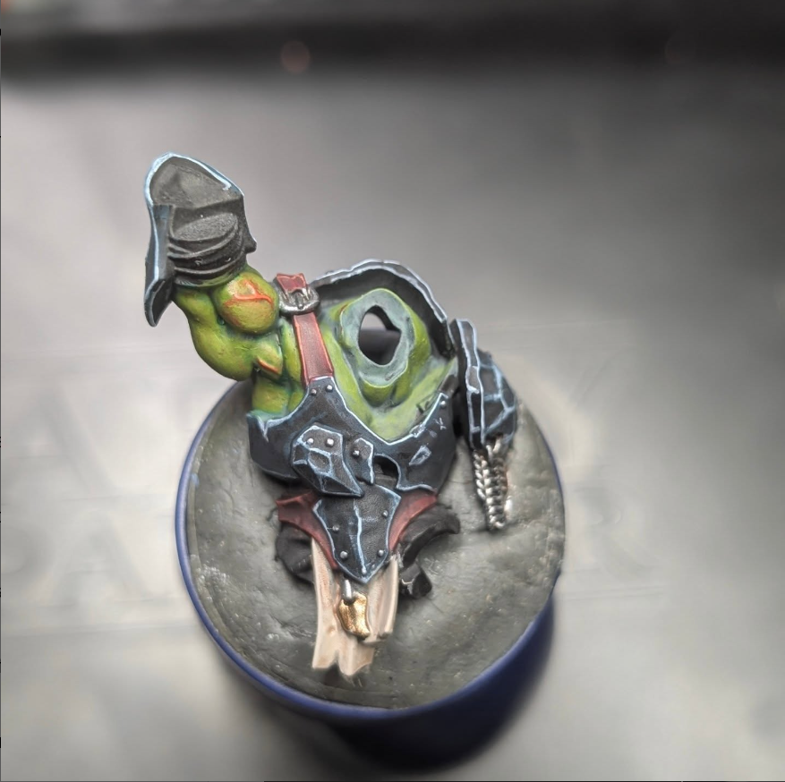 A small sculpted figure of a turtle wearing a black hat and armor, featuring a chain, and painted in green, black, red, and brown, mounted on a circular base.