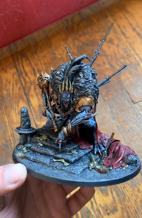 A detailed fantasy miniature figure of a beastly creature with multiple limbs, armor, and weapons, kneeling on a textured base with ruins and bones.