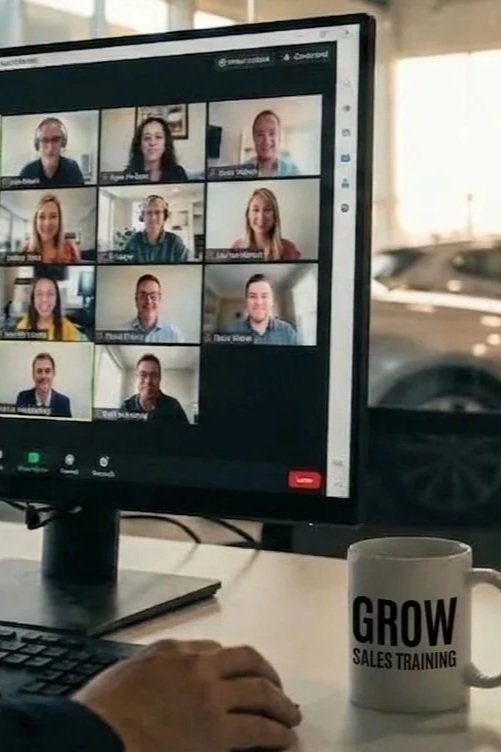 Computer screen displaying a virtual meeting with nine participants. A hand is typing on a keyboard, and a coffee mug labeled 'GROW SALES TRAINING' is on the desk in front of the monitor. By Lambo Borgardt
