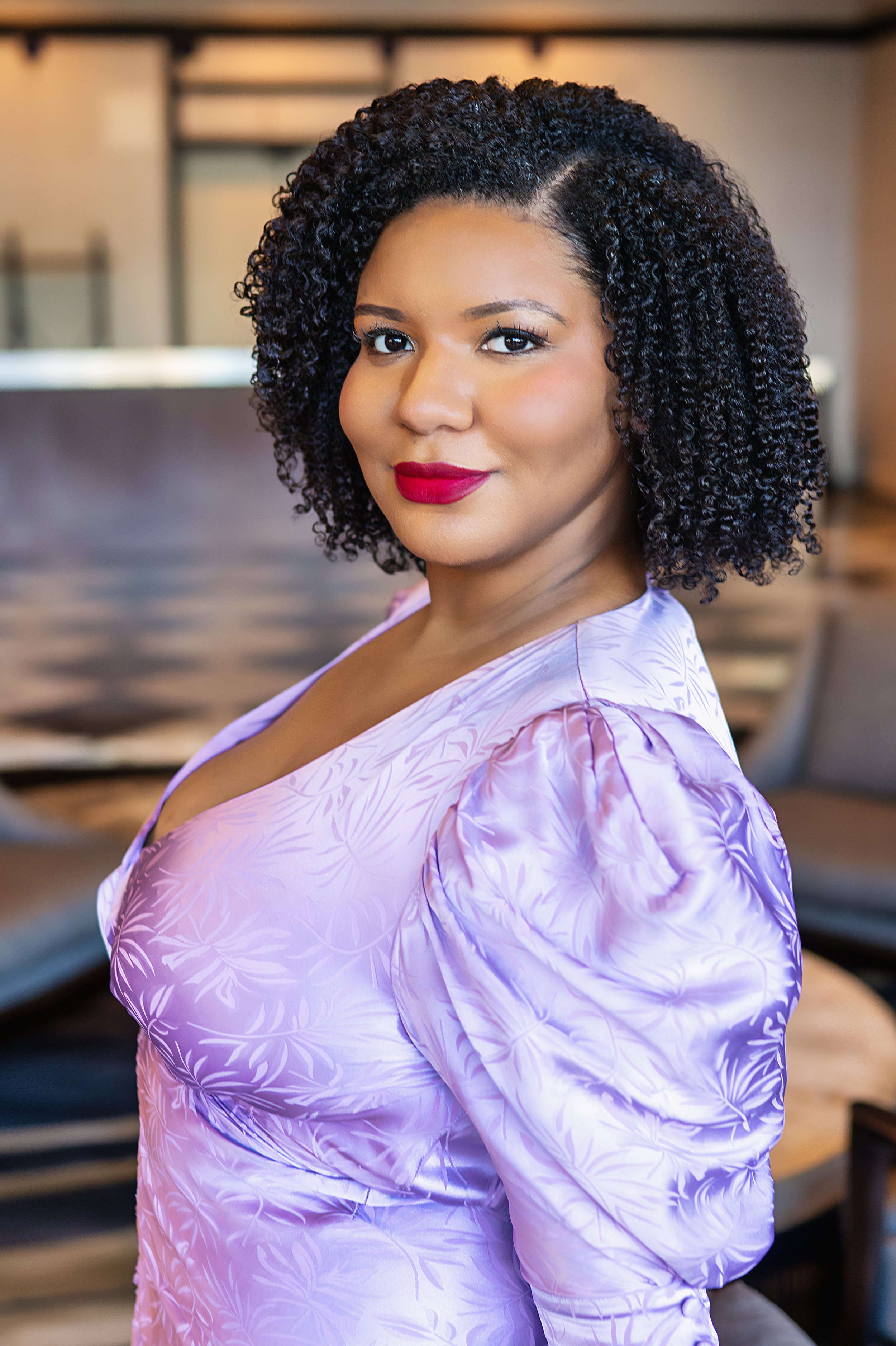 A woman with dark curly hair and makeup, wearing a light purple dress, standing indoors with blurred furniture in the background.