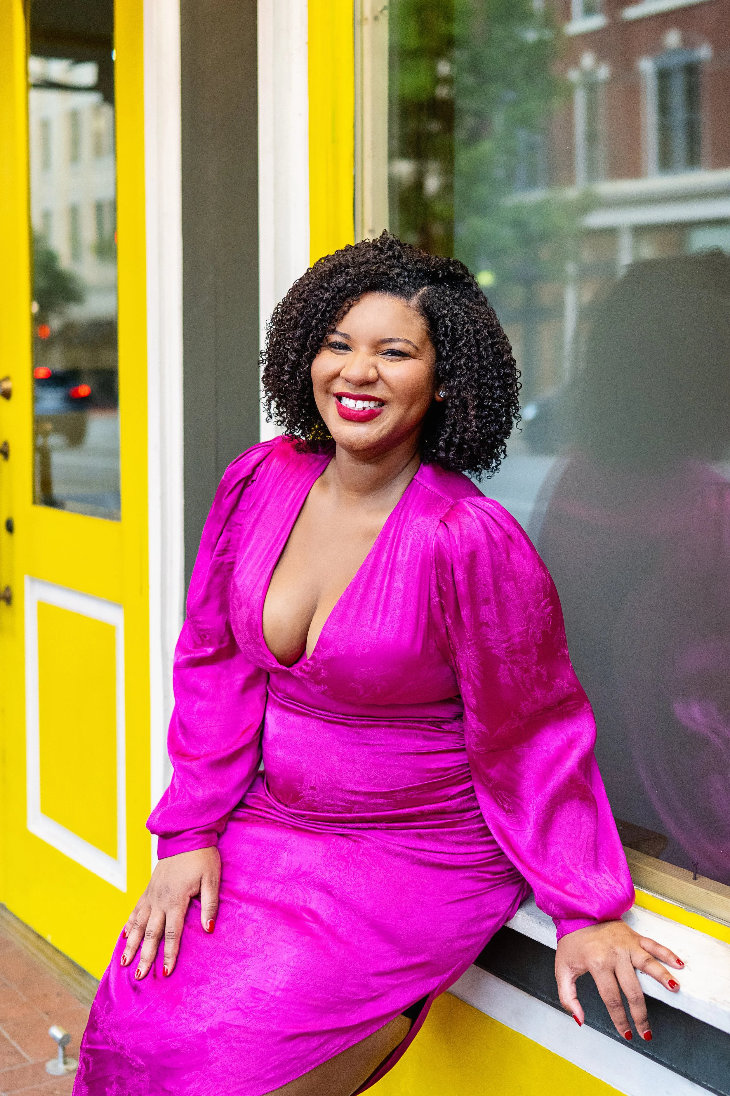 A woman with curly black hair and bright pink lipstick is smiling, wearing a vibrant pink satin dress, sitting on a window ledge outside a yellow shop with a neon yellow door, in an urban setting.