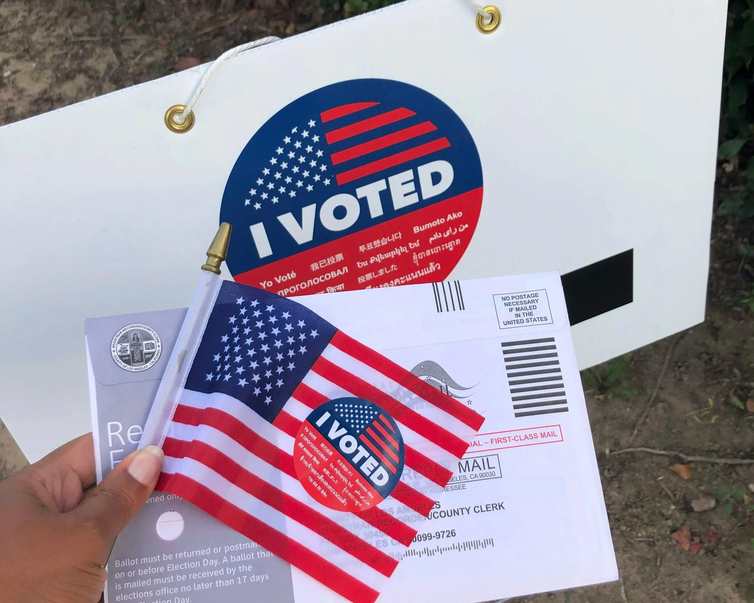 A person holding and pinning a small American flag sticker with an 'I VOTED' sticker on it over a ballot paper, with a large 'I VOTED' sign in the background.