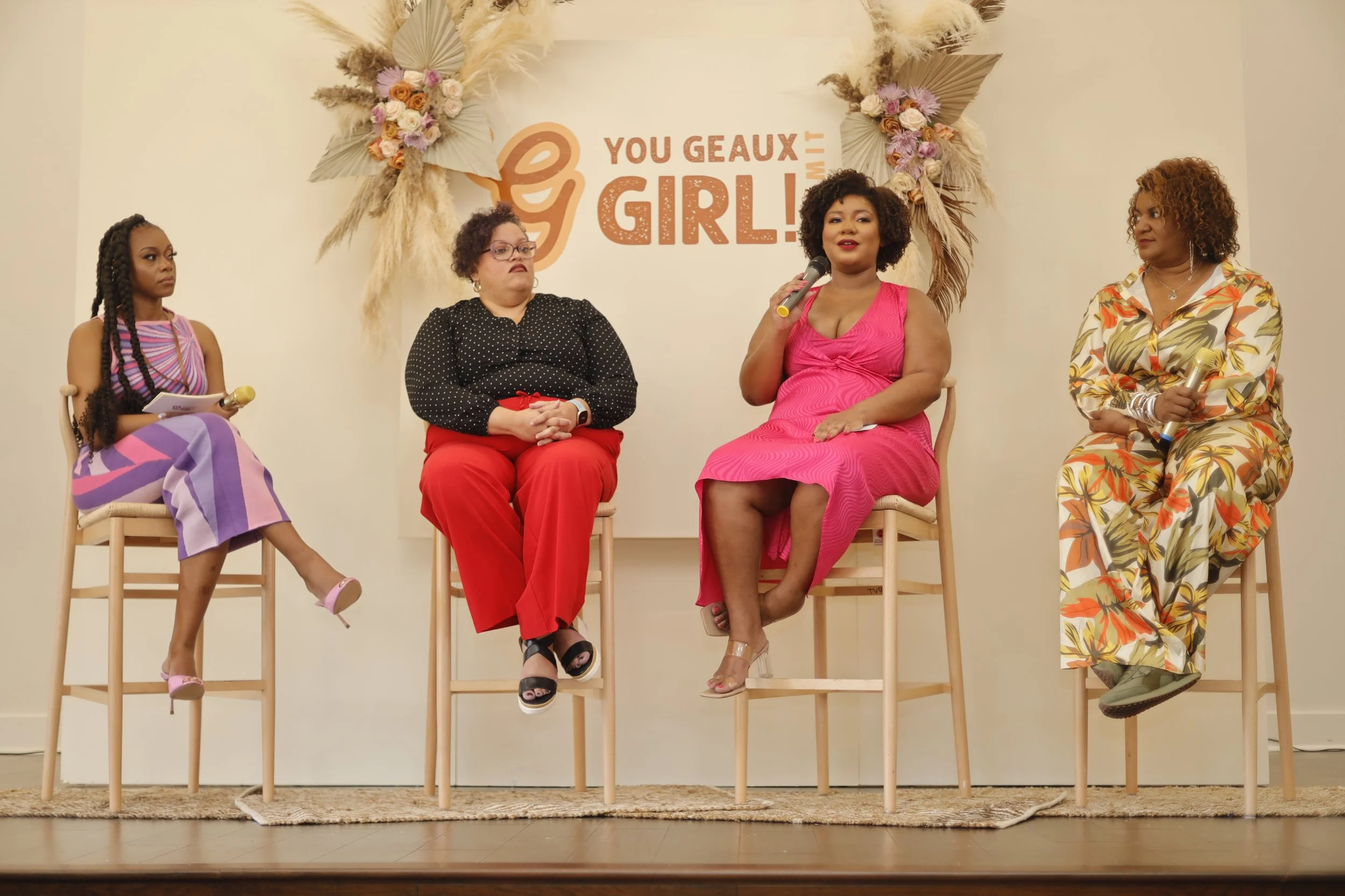 Four women seated on stage during a panel discussion with a floral display backdrop and the words "You Gaux Girl" on the wall behind them.