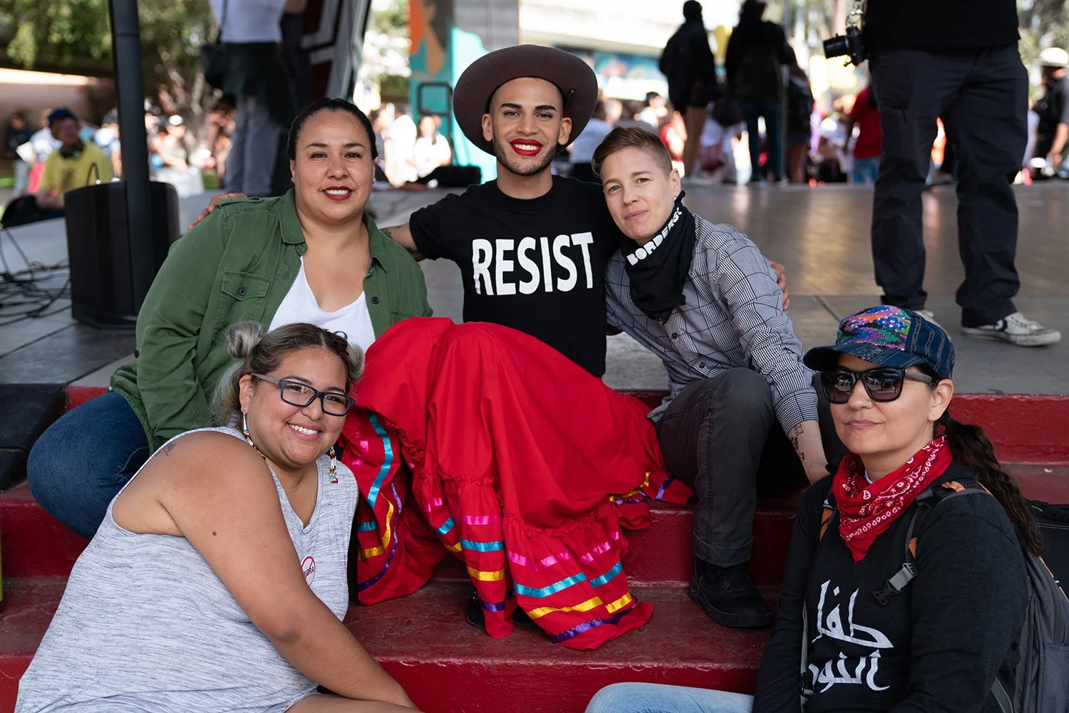 Group of six diverse young women at an outdoor event, sitting and kneeling on red steps, with some smiling at the camera, while a colorful mural and other people are visible in the background.