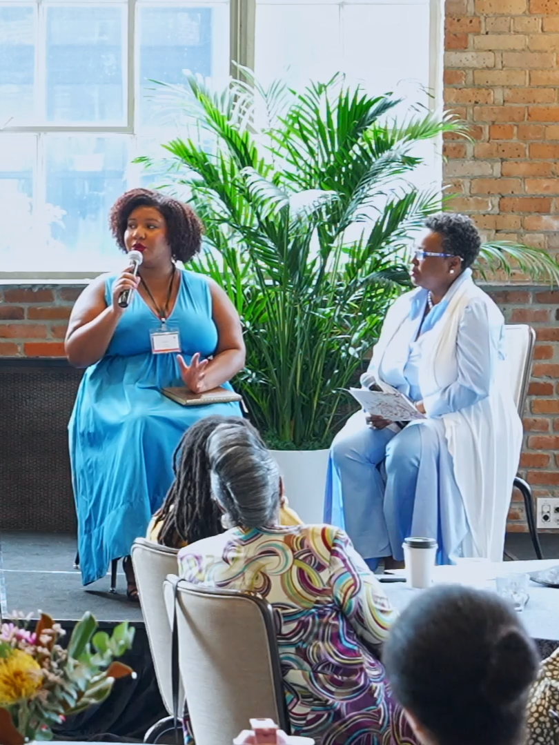 Two women speaking at an event, one holding a microphone and the other holding a paper, seated near a large green plant in a room with brick walls and large windows. Audience members are visible in the foreground.