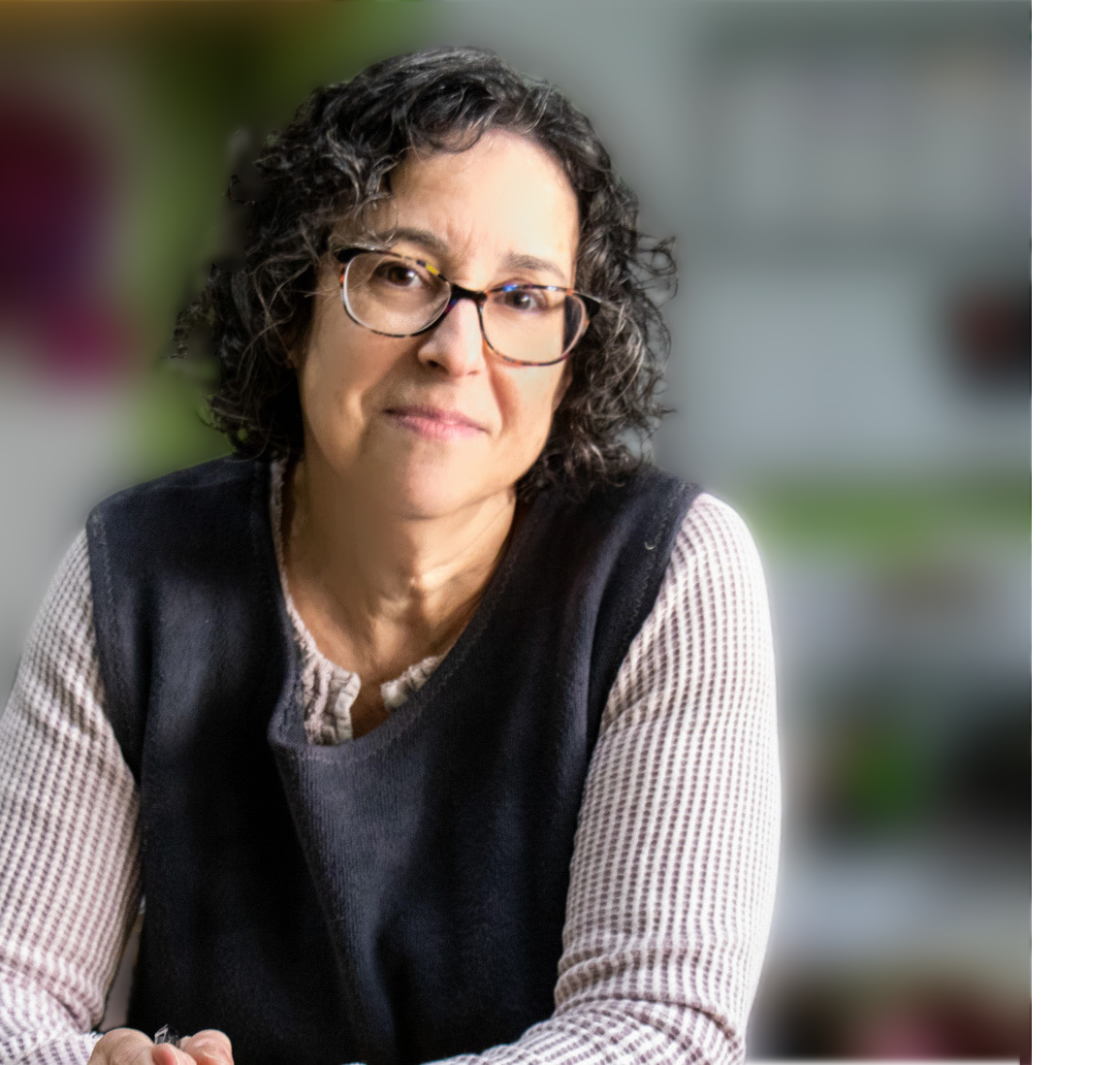 A woman with curly dark hair and glasses looking at the camera with a slight smile, wearing a black vest over a cream-colored long-sleeve shirt, sitting indoors with blurred background.