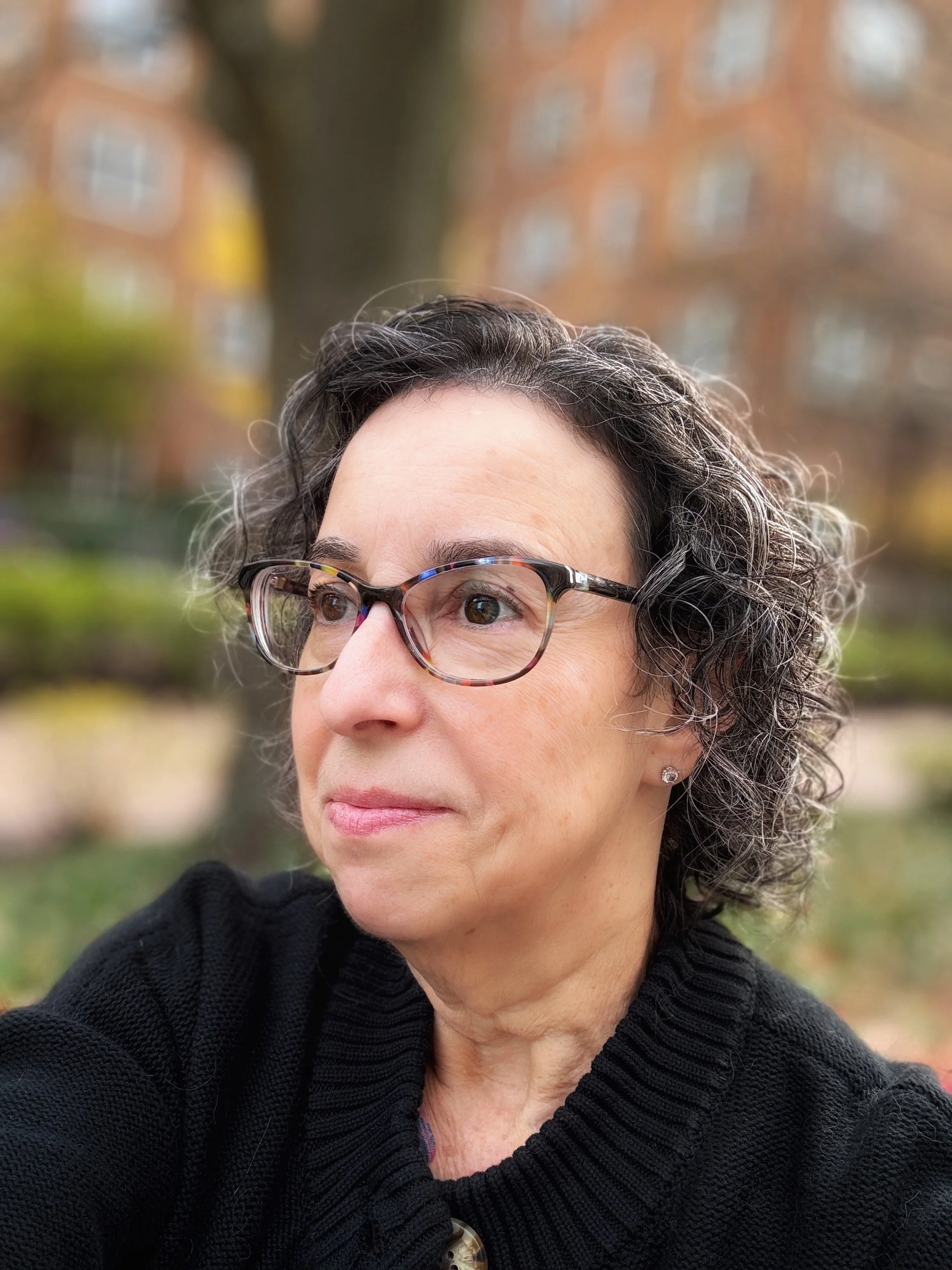 A woman wearing glasses looks to the side outdoors with autumn foliage and buildings blurred in the background.