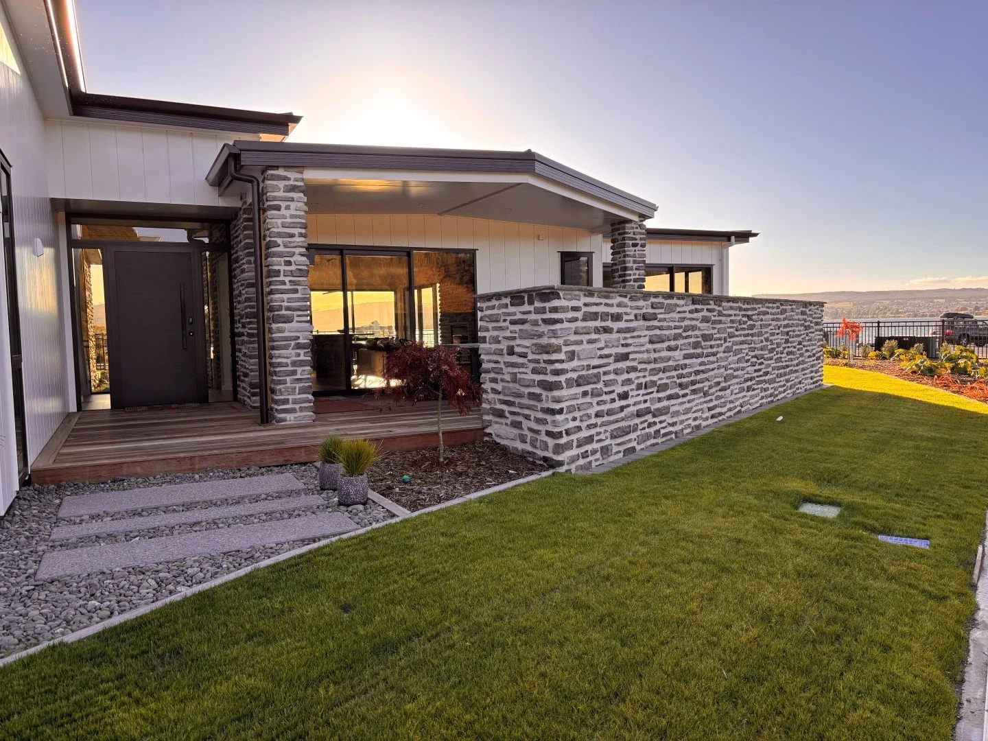 Modern house with stone accent walls and a wooden deck, surrounded by a green lawn and garden in a sunny outdoor setting.